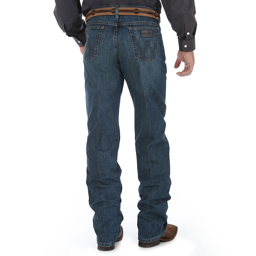 A man wearing Wrangler Men's 20X Competition Jean in a river wash color, sitting with one hand in pocket and looking off to the distance.