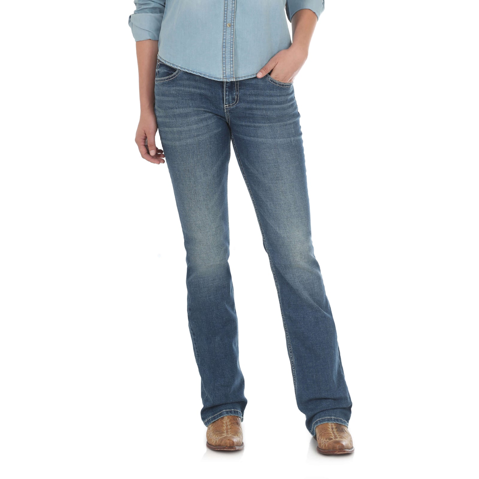 Wrangler Women's Mae Retro Mid Rise Jean. A woman standing and posing in Wrangler women's Mae Retro mid-rise blue jeans with open pockets and 'W' stitching on the back pockets, wearing a chambray shirt and brown boots.