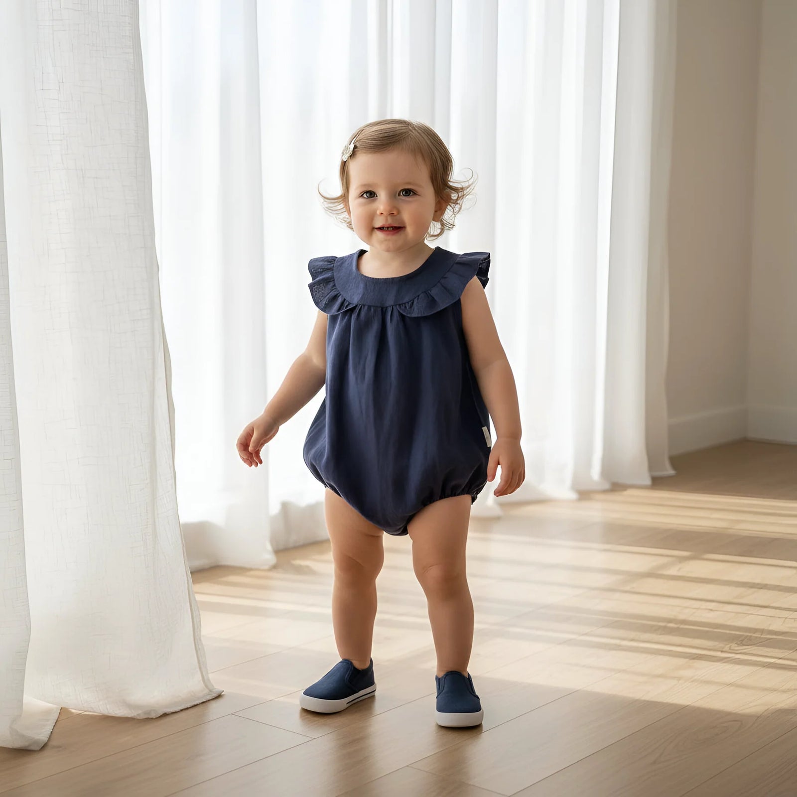 A toddler stands indoors on a wood floor in front of white curtains, wearing a navy sleeveless romper with ruffled shoulders and matching navy shoes.