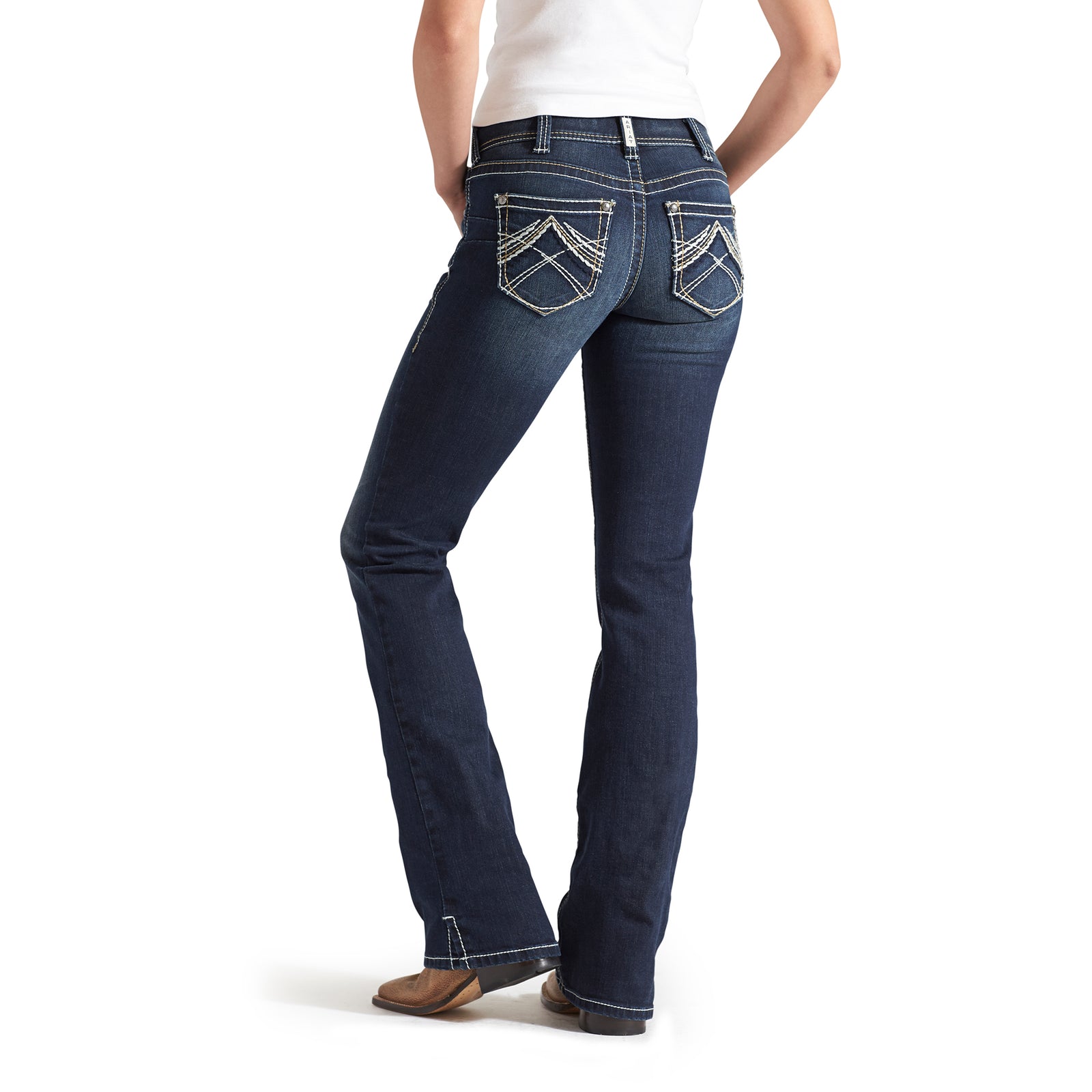 Ariat Women's Real Mid Rise Boot Cut Jean. Person wearing dark blue jeans with hands in pockets on a white background