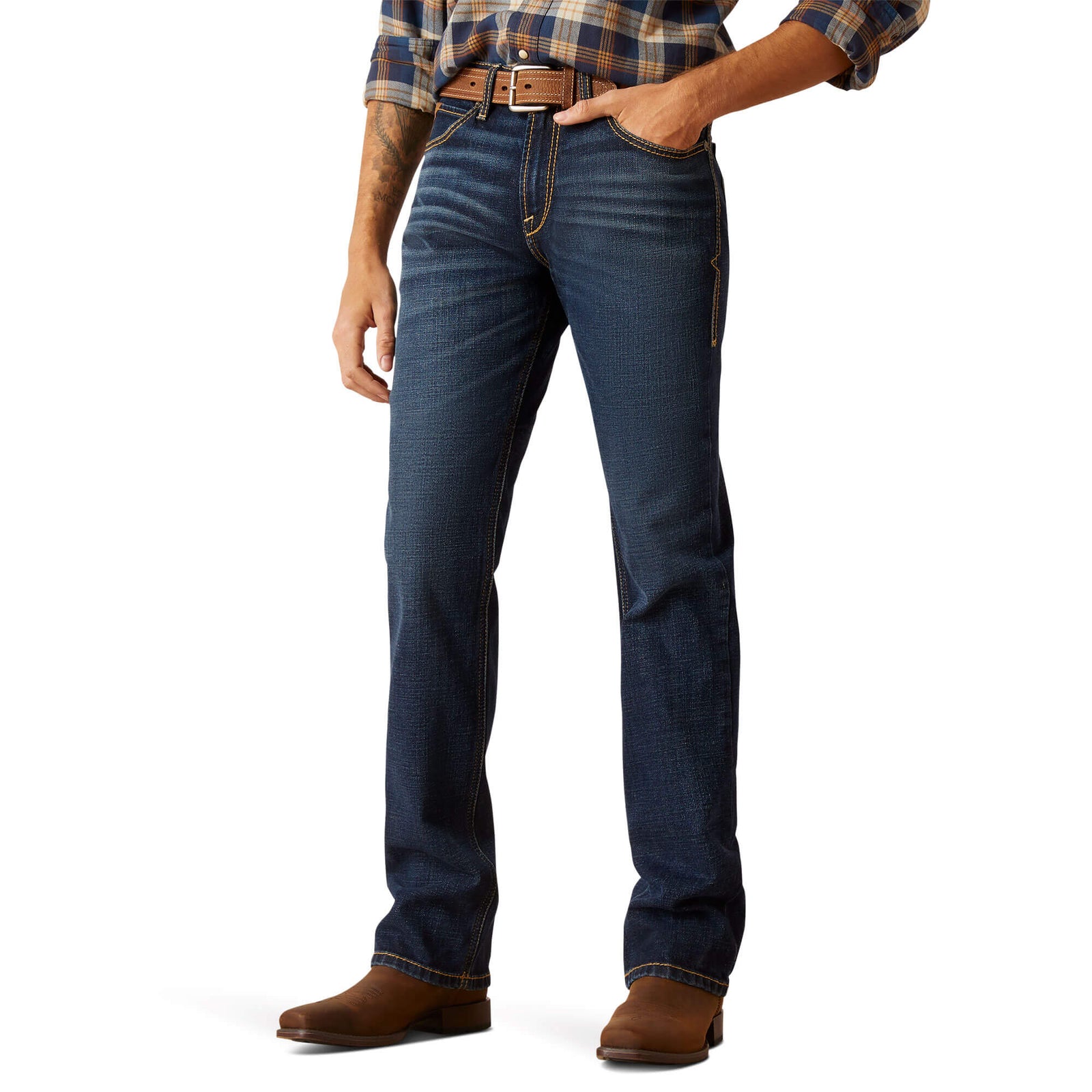 A man standing wearing Ariat Men's M8 Reese Modern Fit Jeans and brown boots. The jeans are blue with a subtle pattern, and the man has his hands in his pockets.