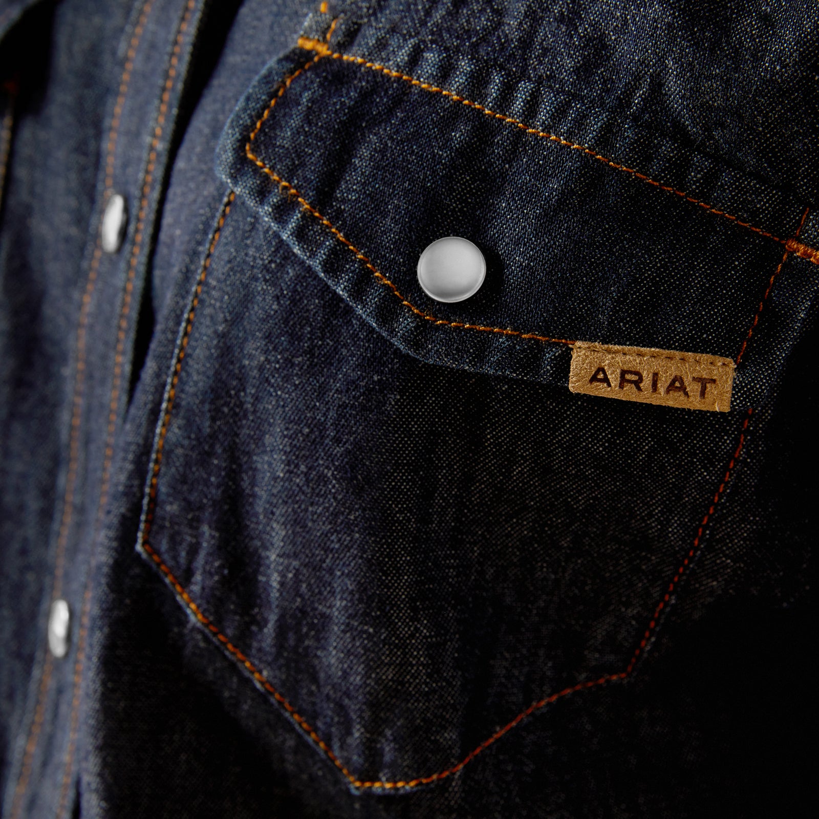 Ariat Women's Farriday Denim  L/S Western Shirt. Close-up of the chest pocket of a blue denim Ariat women's shirt with a visible brand tag.