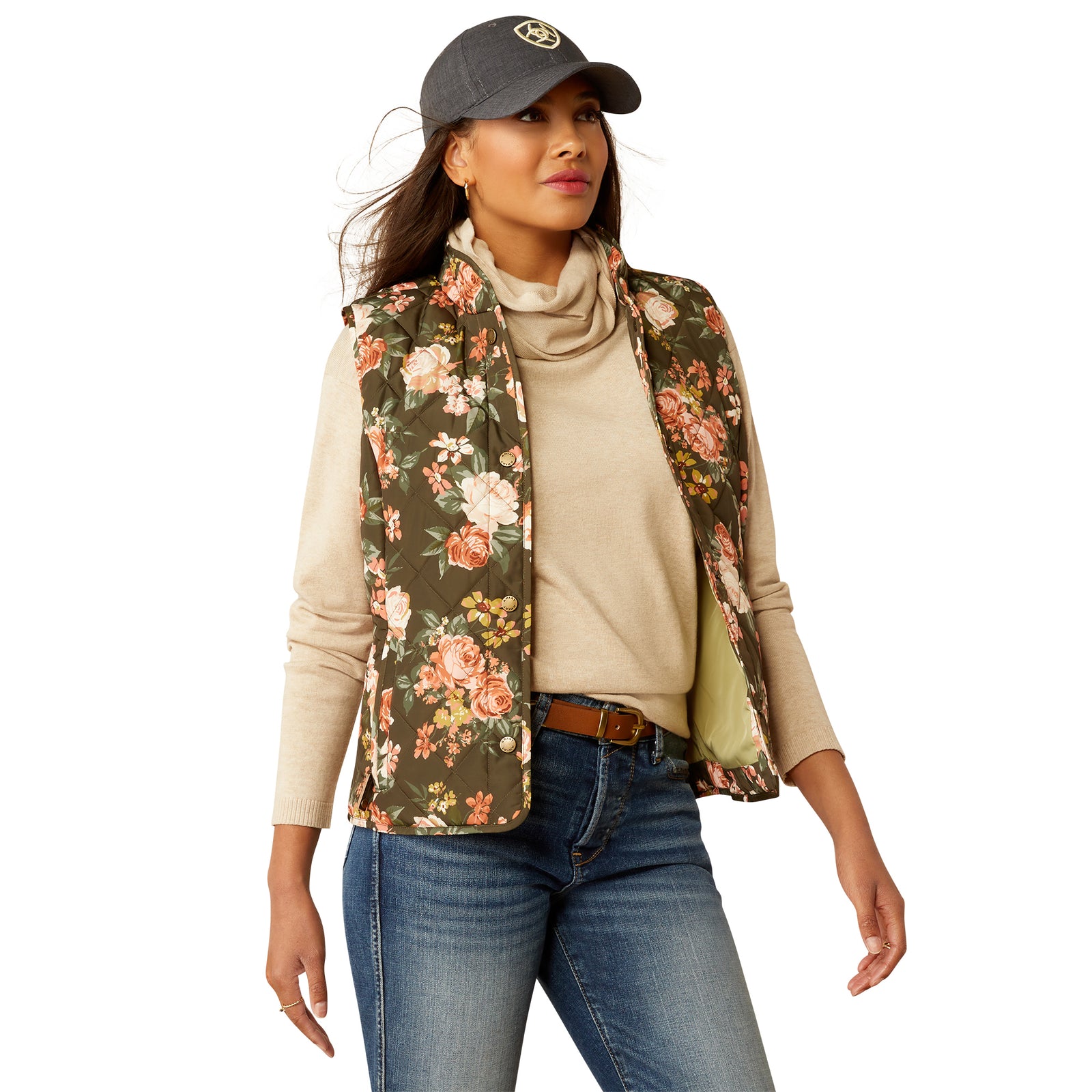Ariat Women's Woodside Vest. A woman wearing a floral quilted Ariat vest with a turtleneck sweater, jeans, and a cap.