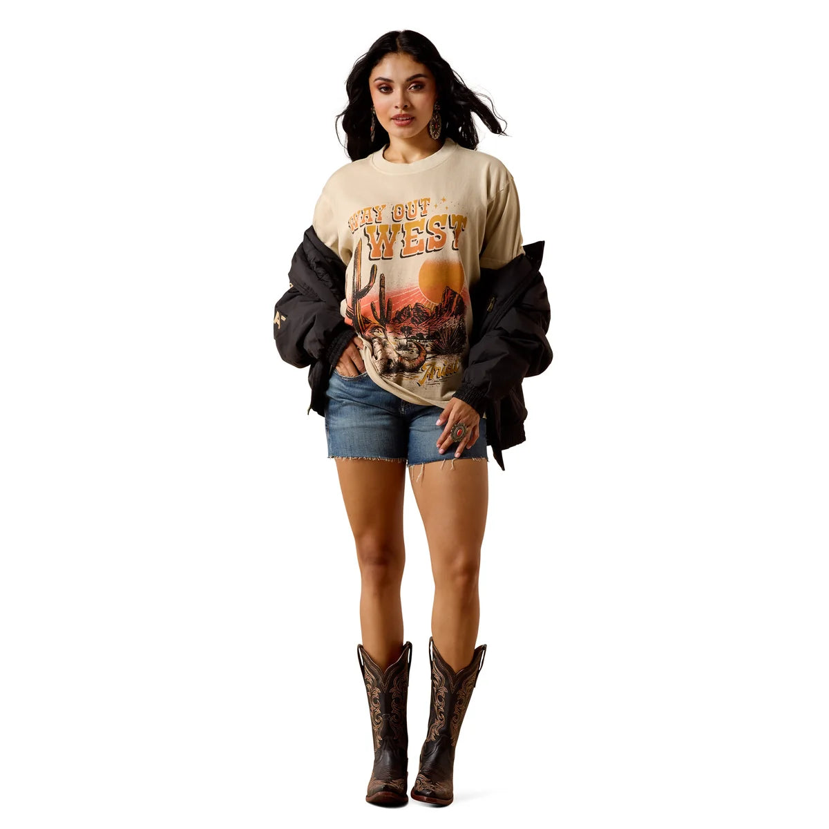 A woman modeling a beige graphic t-shirt with 'Say Out West' and a cowboy illustration, styled with denim shorts, a black jacket draped off her shoulders, and brown cowboy boots against a white background.