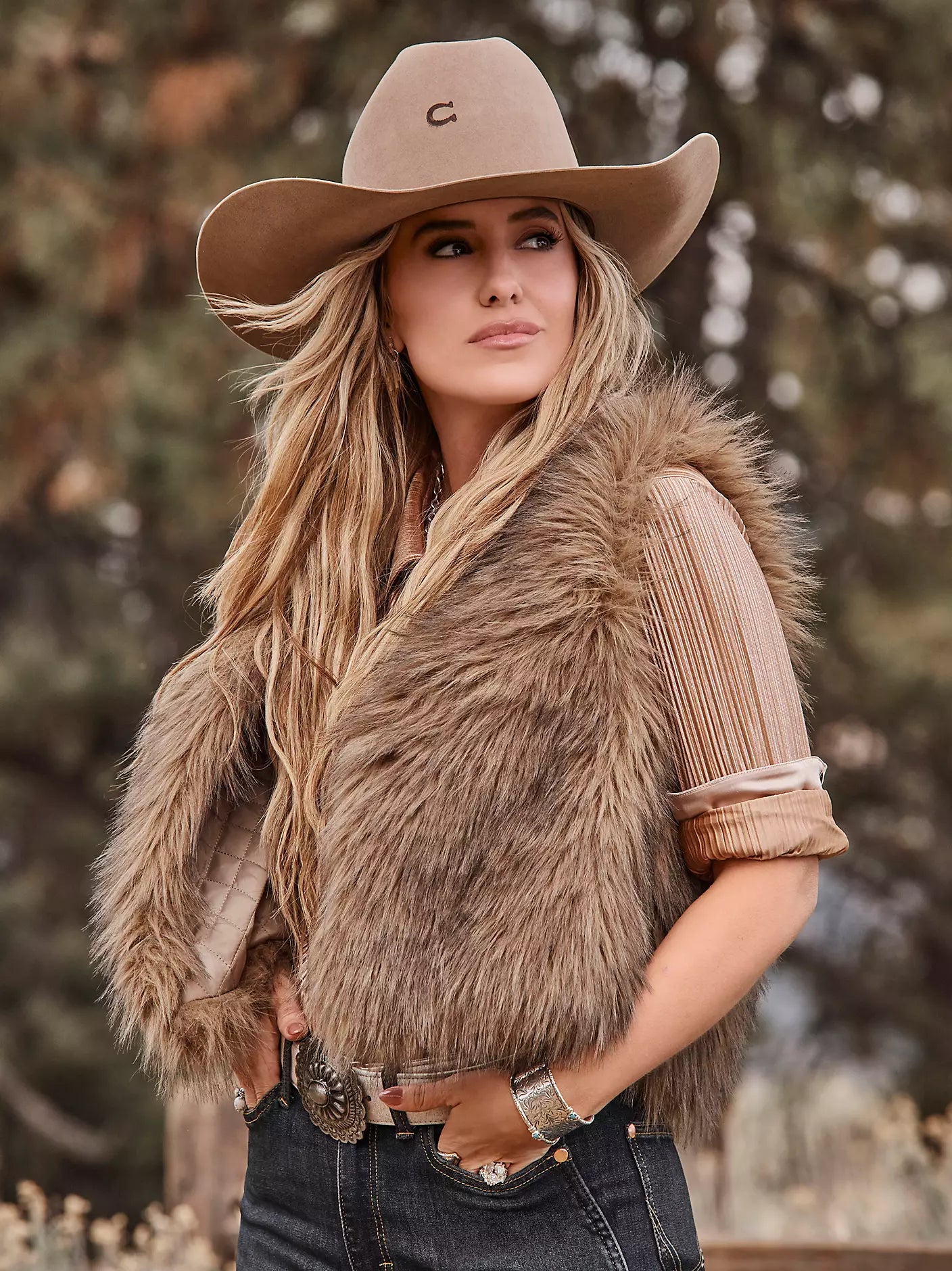 A woman wearing a tan cowboy hat with a small embroidered 'C' logo, a brown faux fur vest over a beige pleated top, and dark blue jeans, posed outdoors against a blurred forest background.