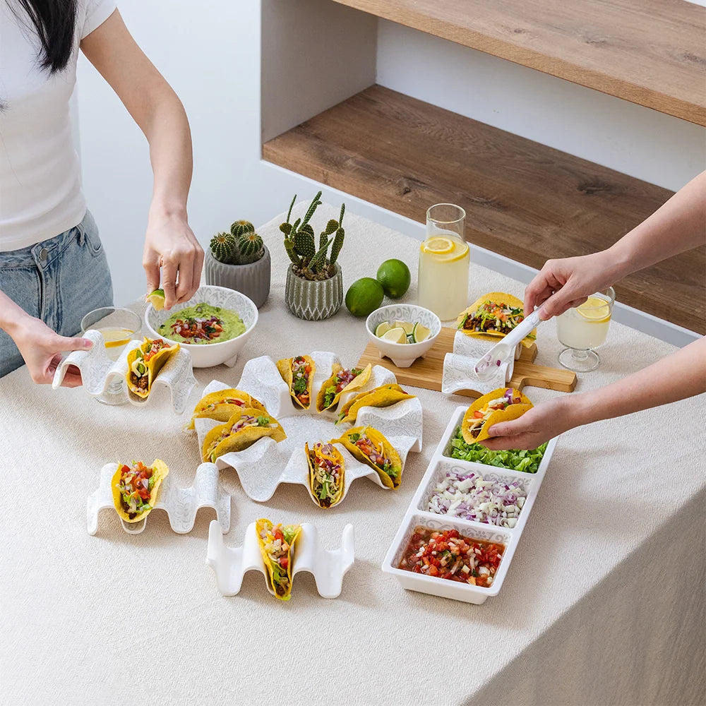 White ceramic taco holder set arranged on a dining table, with people serving and holding tacos in a bright indoor setting. The set includes multiple taco stands, a three-compartment dipping tray, and small bowls of salsa and guacamole.