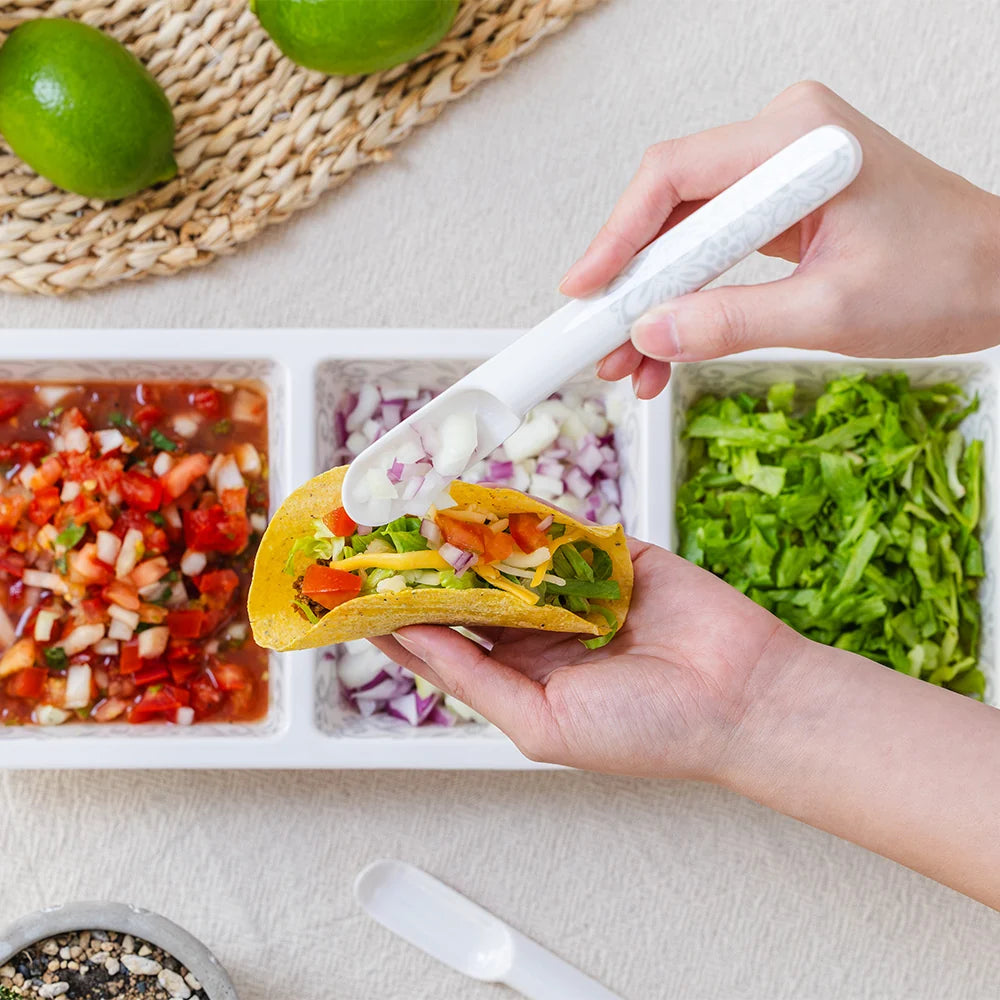 A close-up of a person using a white plastic taco filling spreader to scoop sour cream onto a taco, with a three-compartment tray of ingredients and limes in the background.