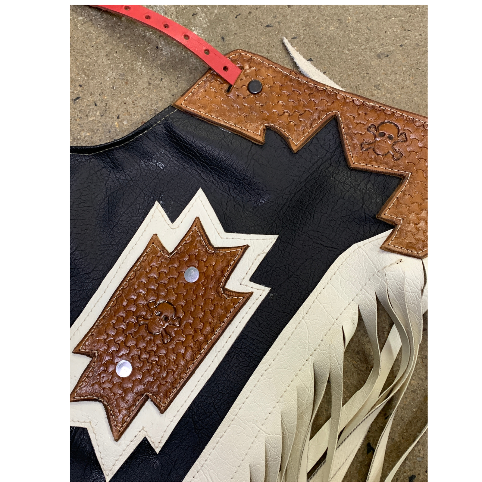 A close-up of a western-style leather chaps with black body, tan and white geometric trim, fringe, and a red belt loop, photographed on a concrete surface.