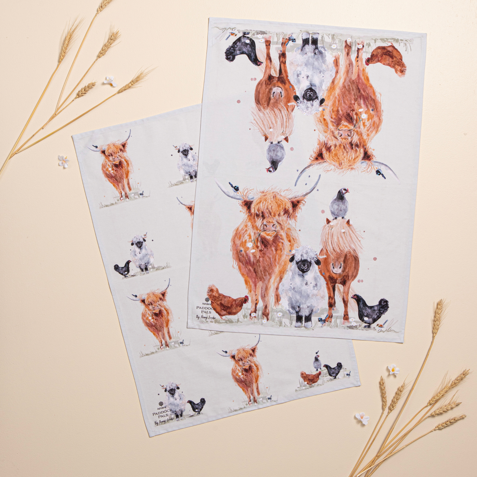 Two white tea towels with a whimsical watercolor illustration of Highland cows, sheep, chickens, and ducks, arranged in a flat-lay on a beige background with decorative wheat sprigs.
