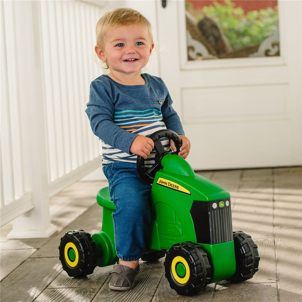 John Deere Foot to Floor - Tractor Ride-On. Child sitting on a green toy tractor with John Deere logo indoors.