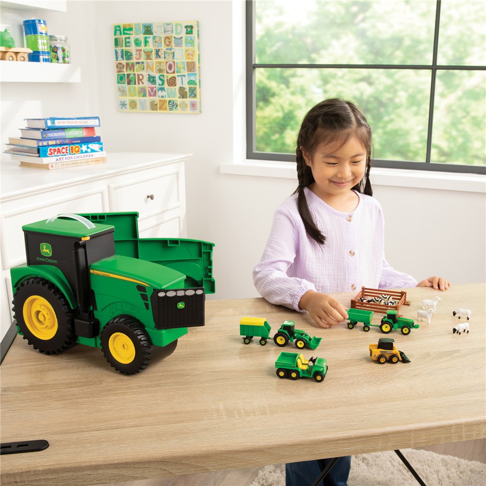 John Deere Carry Case Value Set. Child playing with toy tractors and vehicles on a table in a room with books and a window.