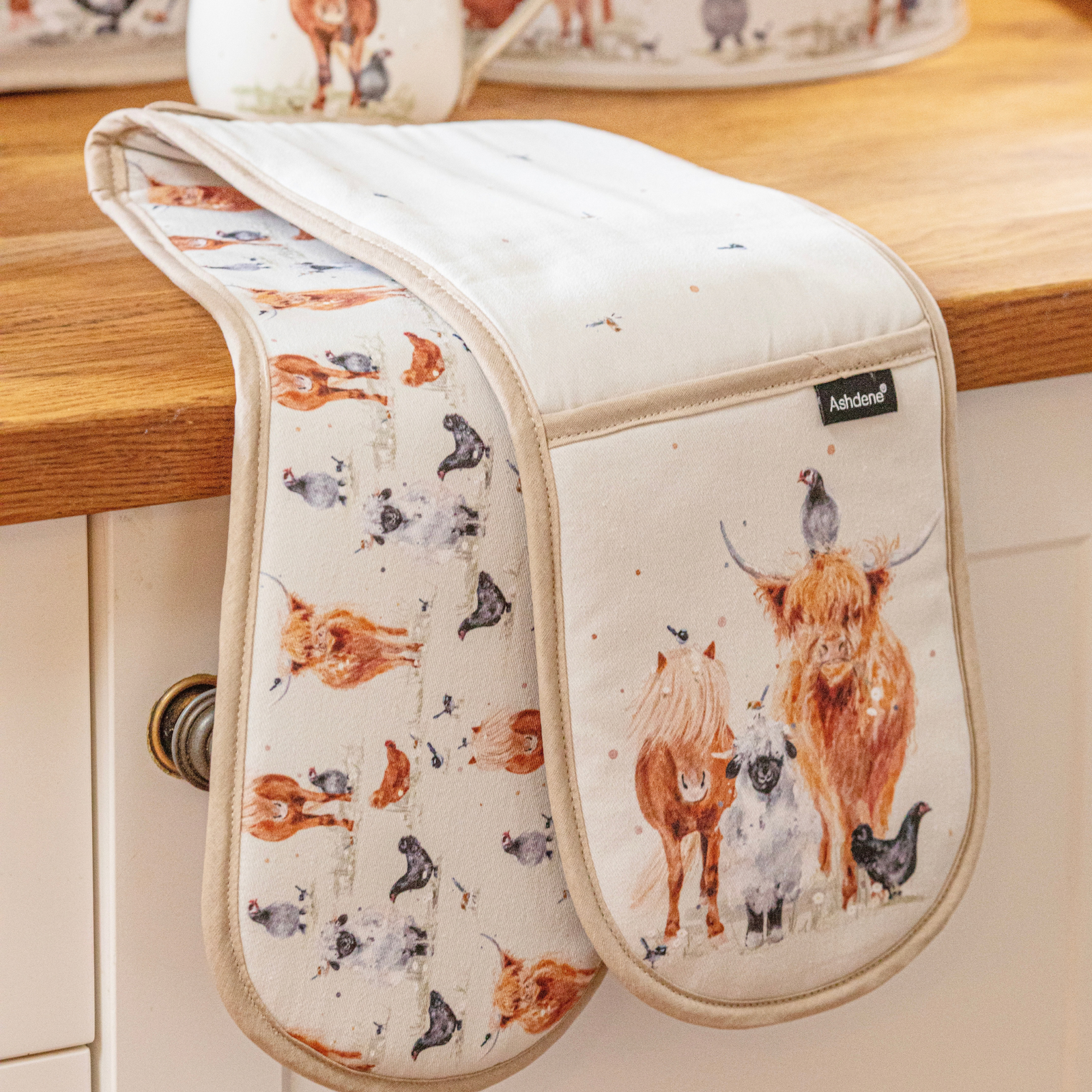 A kitchen oven mitt with a watercolor-style animal print featuring Highland cows, sheep, chickens, and birds, draped over a wooden countertop. The brand name 'Ashand' is visible on a small black label on the front pocket.