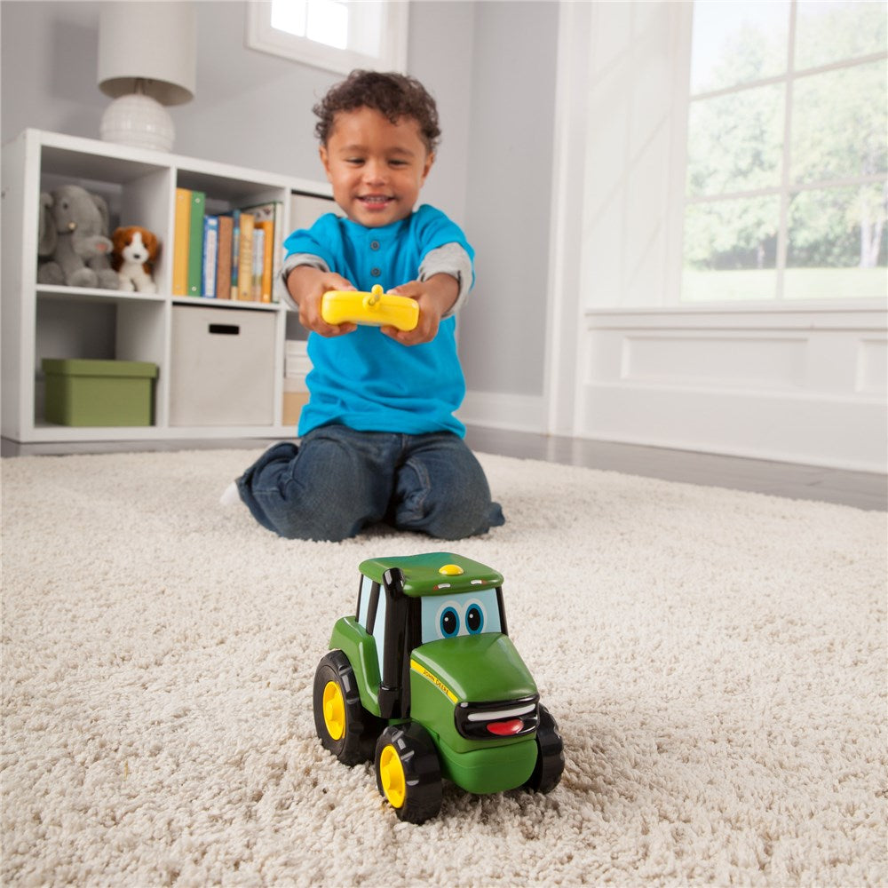 A young child kneeling on a white shag carpet in a bright room, holding a yellow remote control while looking at a green cartoon-style toy tractor with yellow wheels.