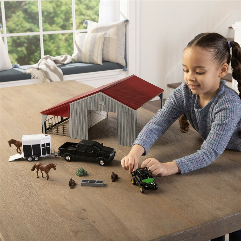 John Deere 1:32 Weathered Barn Play Set. Child playing with toy vehicles and animals on a table with a toy barn in the background.