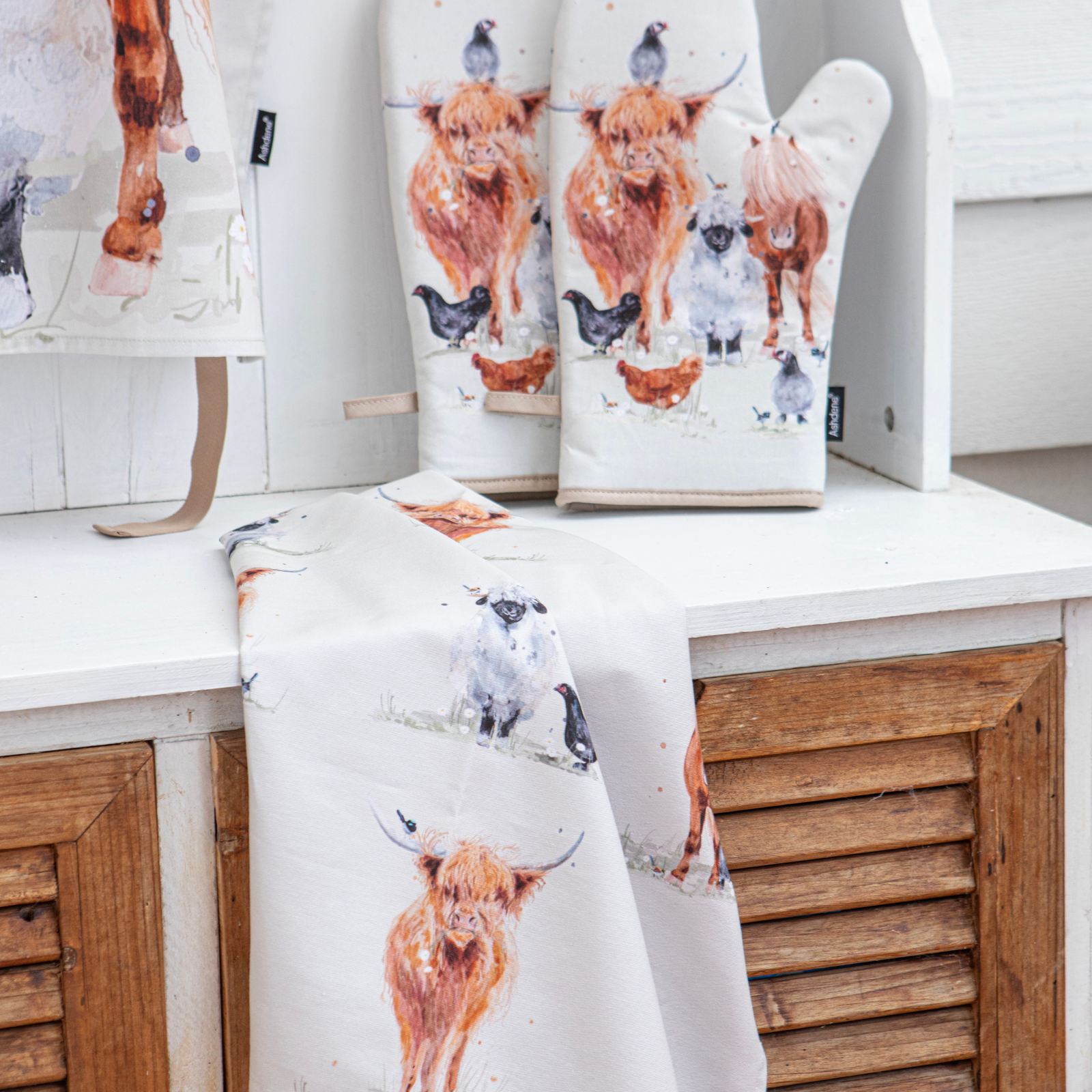 A white kitchen textile set featuring a watercolor-style print of Highland cows, sheep, chickens, and birds, displayed on a white shelf with wooden louvered cabinets behind it. The set includes a towel or cloth draped over the front and matching oven mitts and a hanging cloth behind.