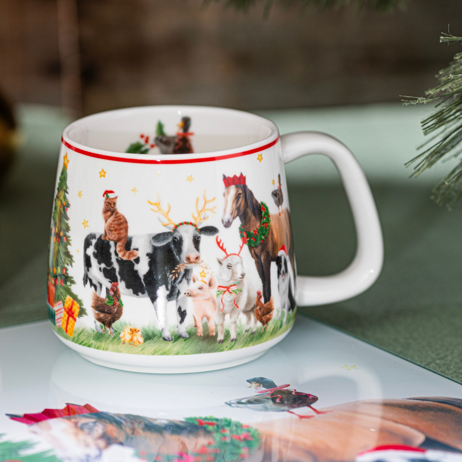 A white ceramic Christmas mug decorated with festive illustrations of a black-and-white cow, a brown horse, a piglet, chickens, a cat, and a Christmas tree, set on a reflective surface.