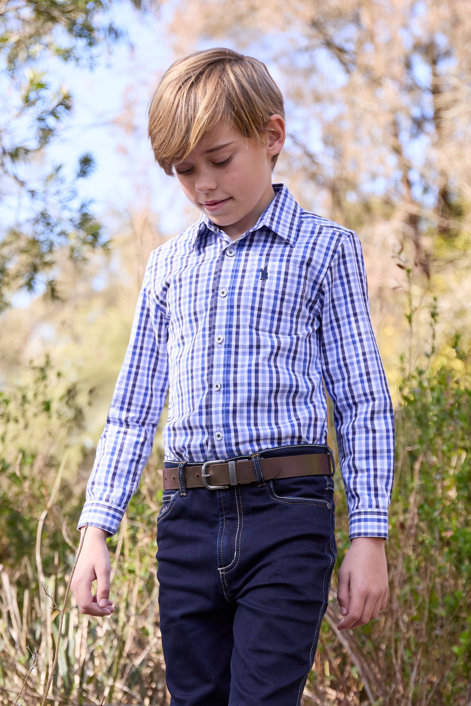 Young boy wearing a blue checkered shirt and dark pants outdoors.