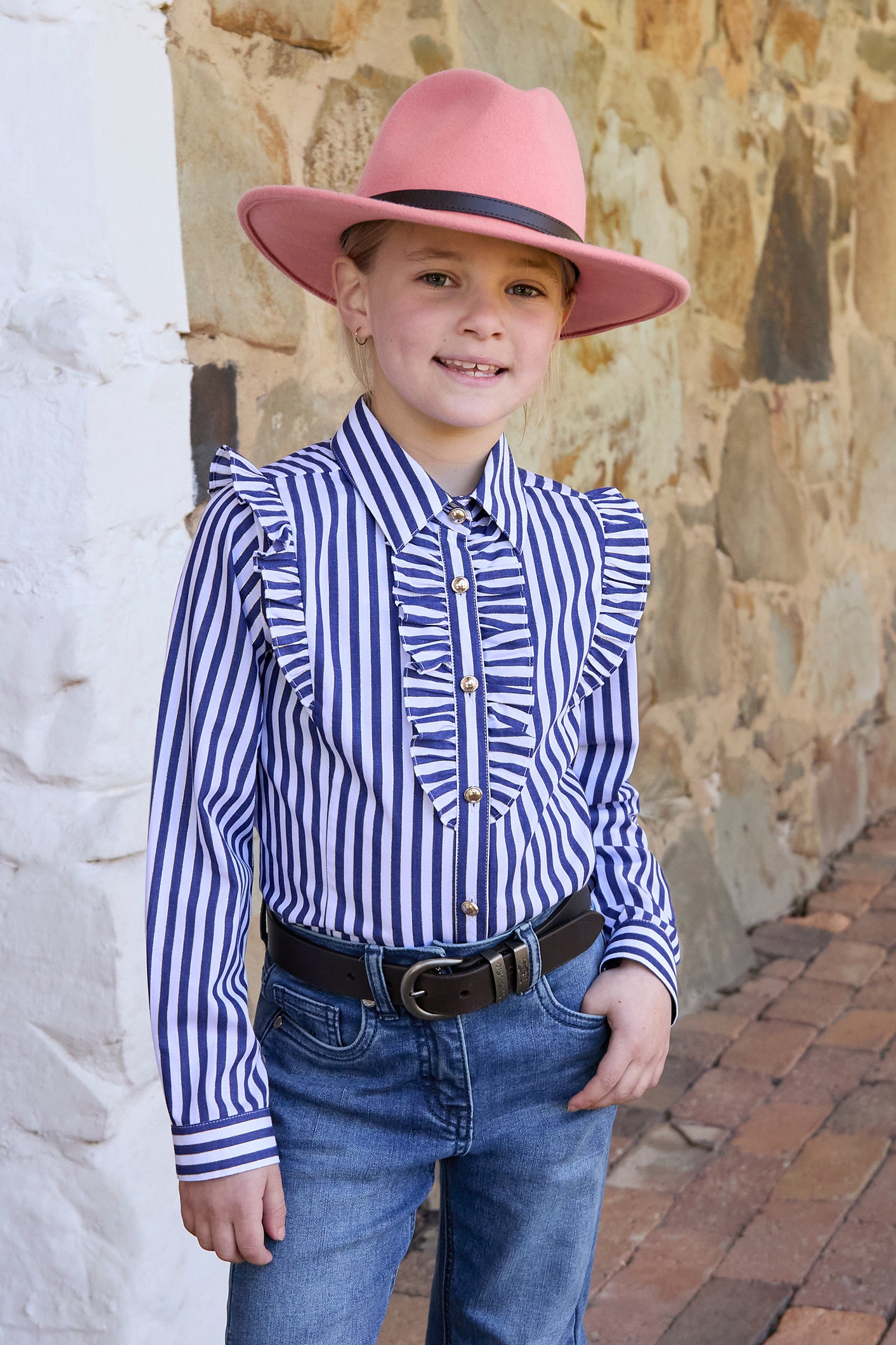 Child wearing a pink hat, striped shirt, and jeans standing against a stone wall.