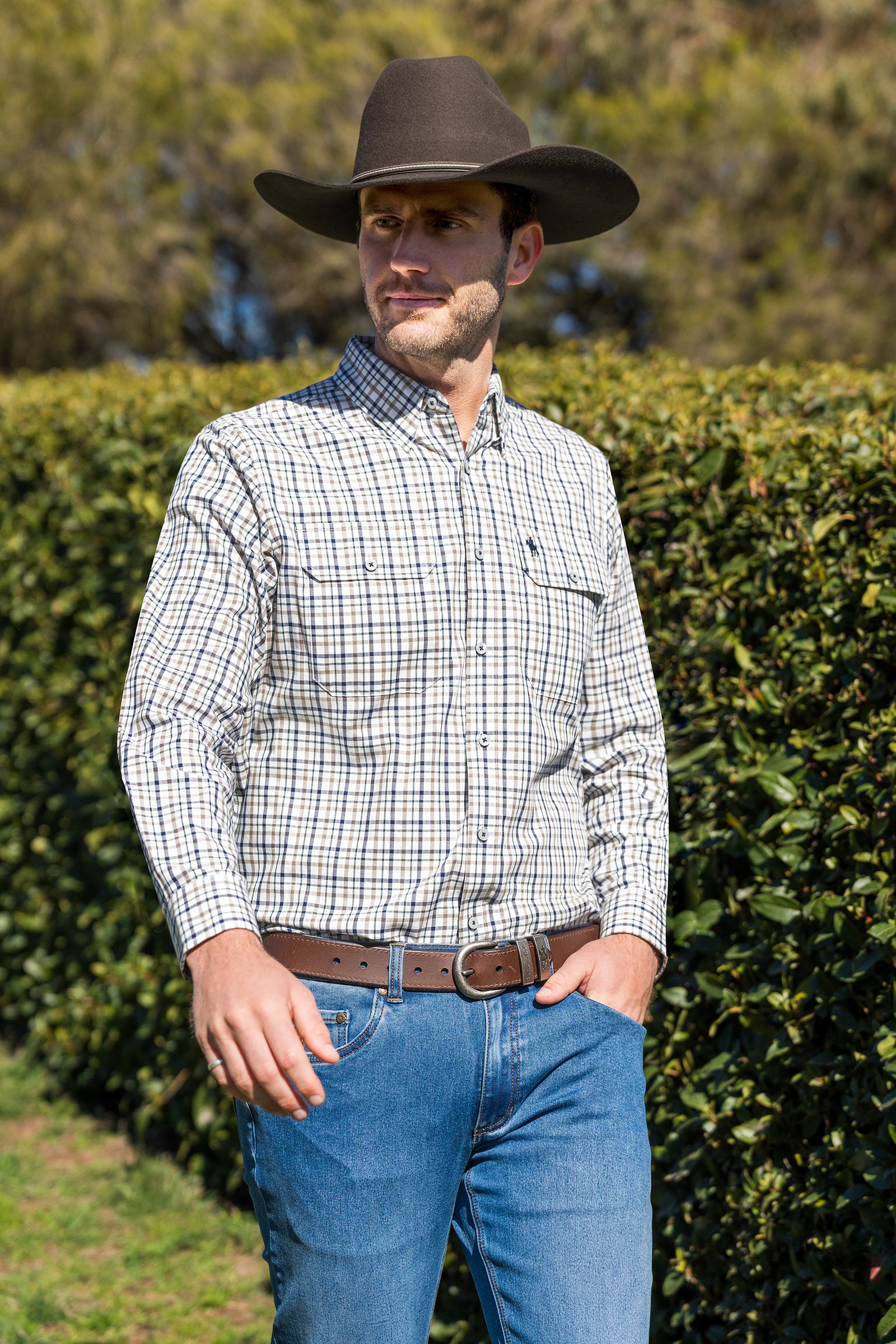 Man wearing a plaid shirt, cowboy hat, and jeans standing in front of green bushes.