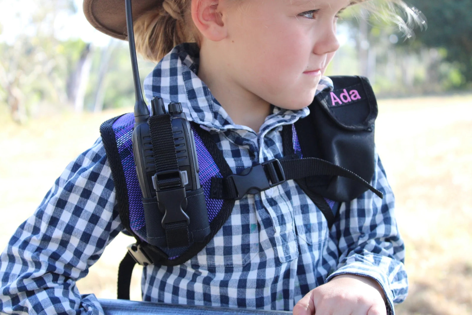 A child wearing a black and purple harness with a mounted walkie-talkie and a black pouch embroidered with the name 'Ada' in pink, outdoors in a sunny field.