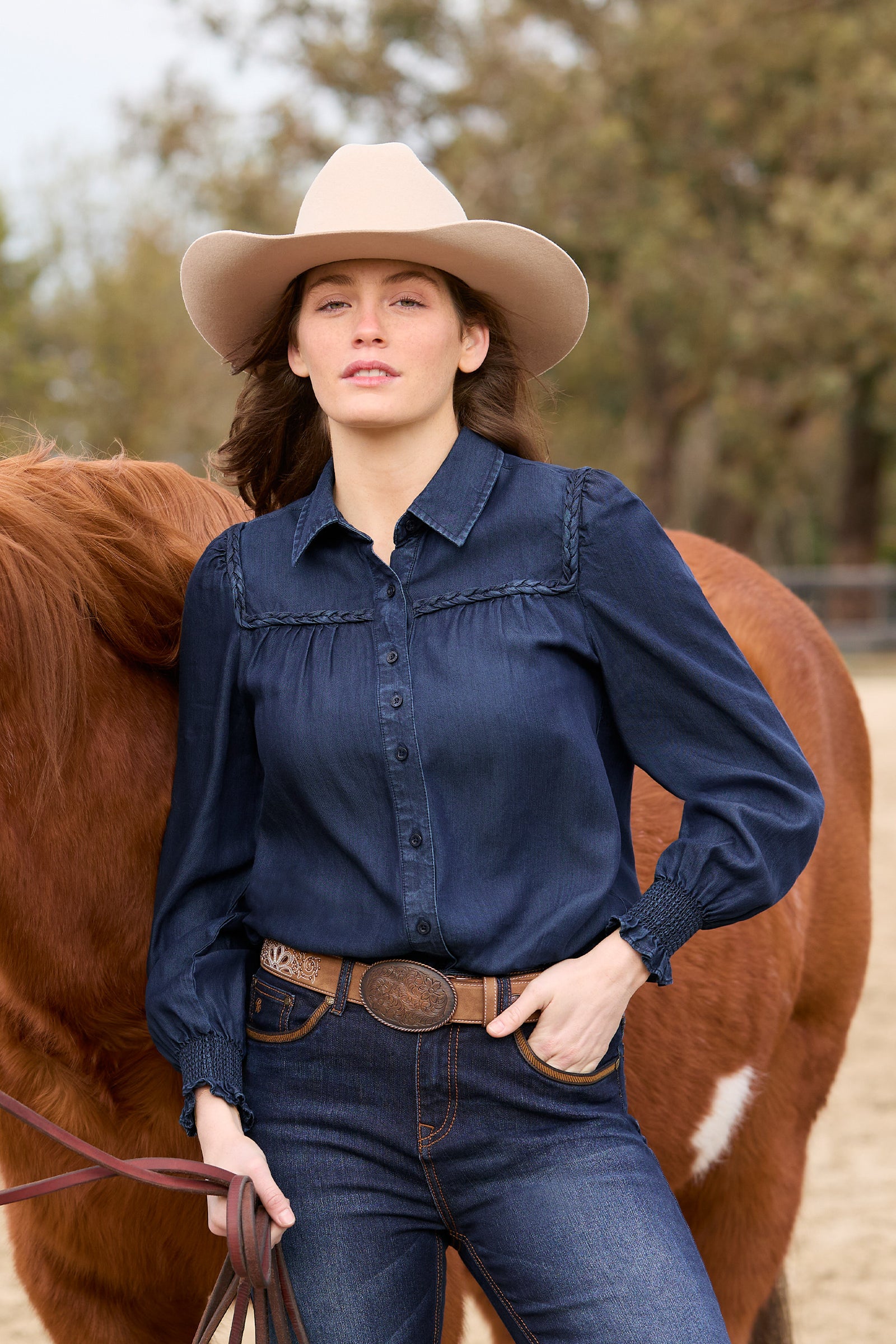 Woman in a blue shirt and cowboy hat standing next to a brown horse in an outdoor setting.