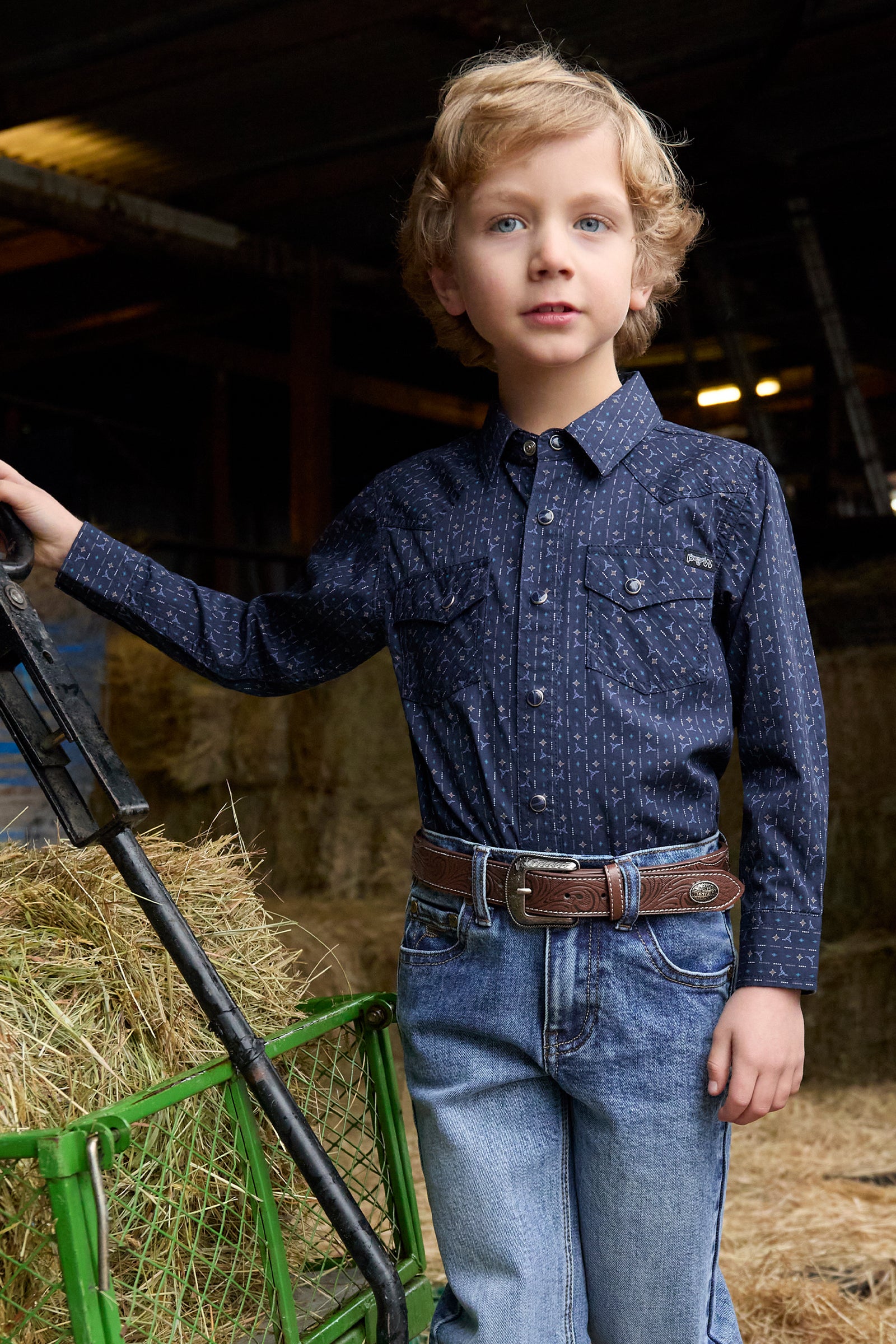 Young boy in a blue shirt and jeans standing in a barn with hay.