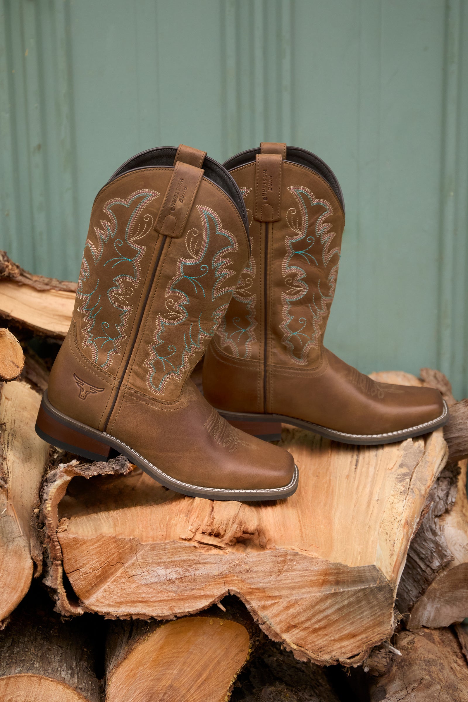 Pair of brown cowboy boots with intricate designs on a stack of wooden logs against a green metal background.