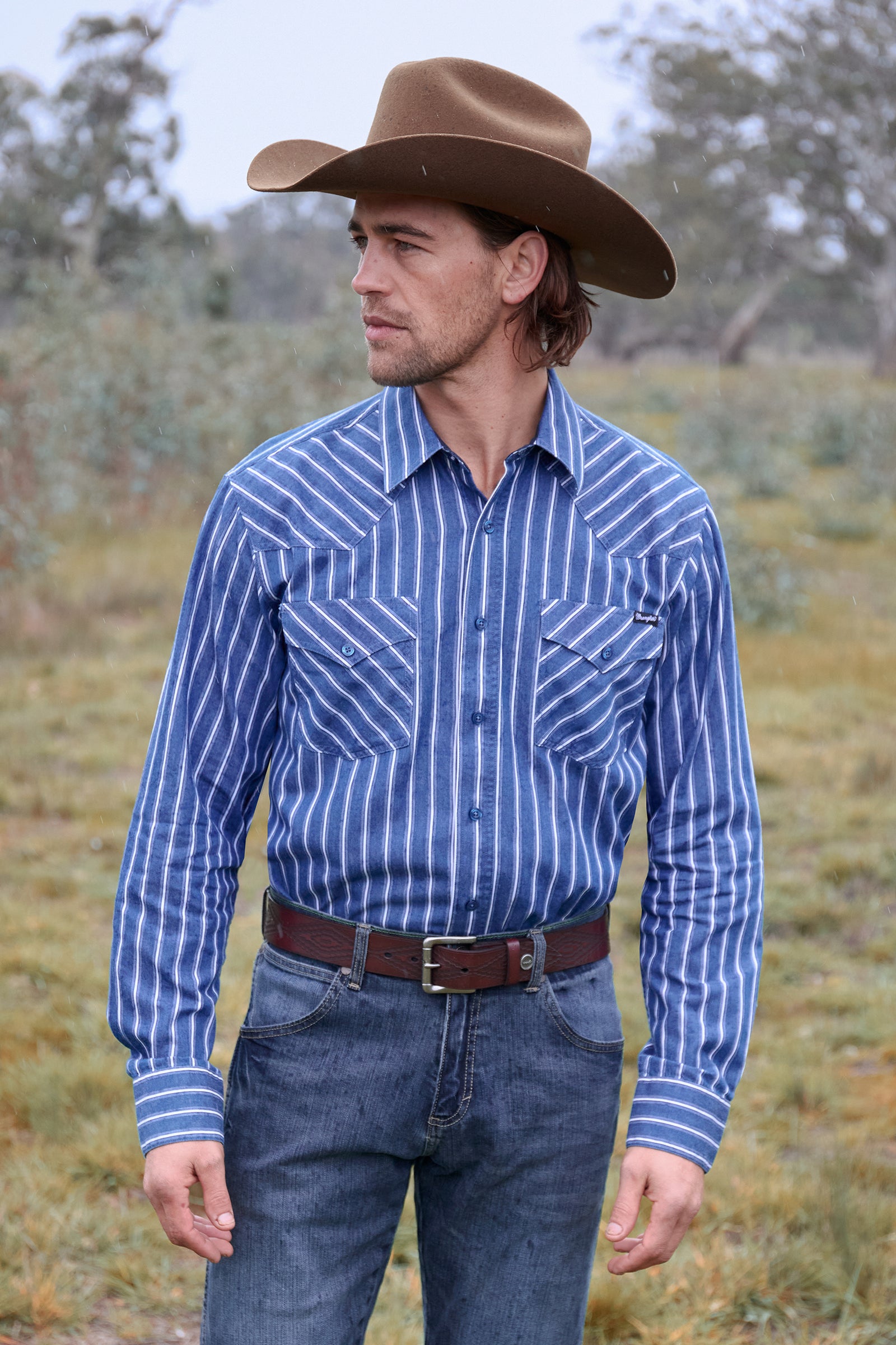 Man wearing a blue striped shirt, brown cowboy hat, and jeans standing in a grassy field.