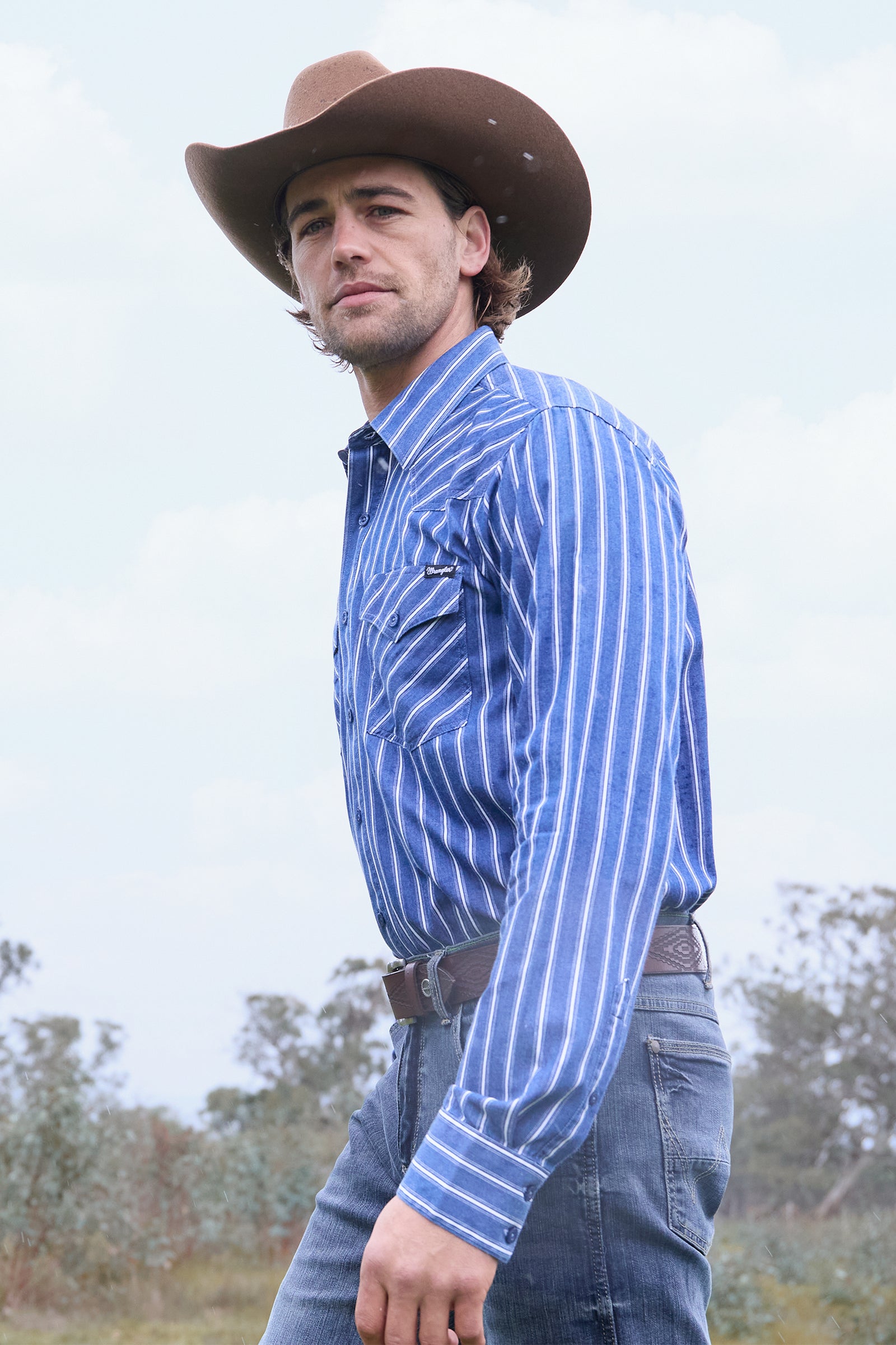 Man wearing a blue striped shirt and cowboy hat outdoors with trees in the background