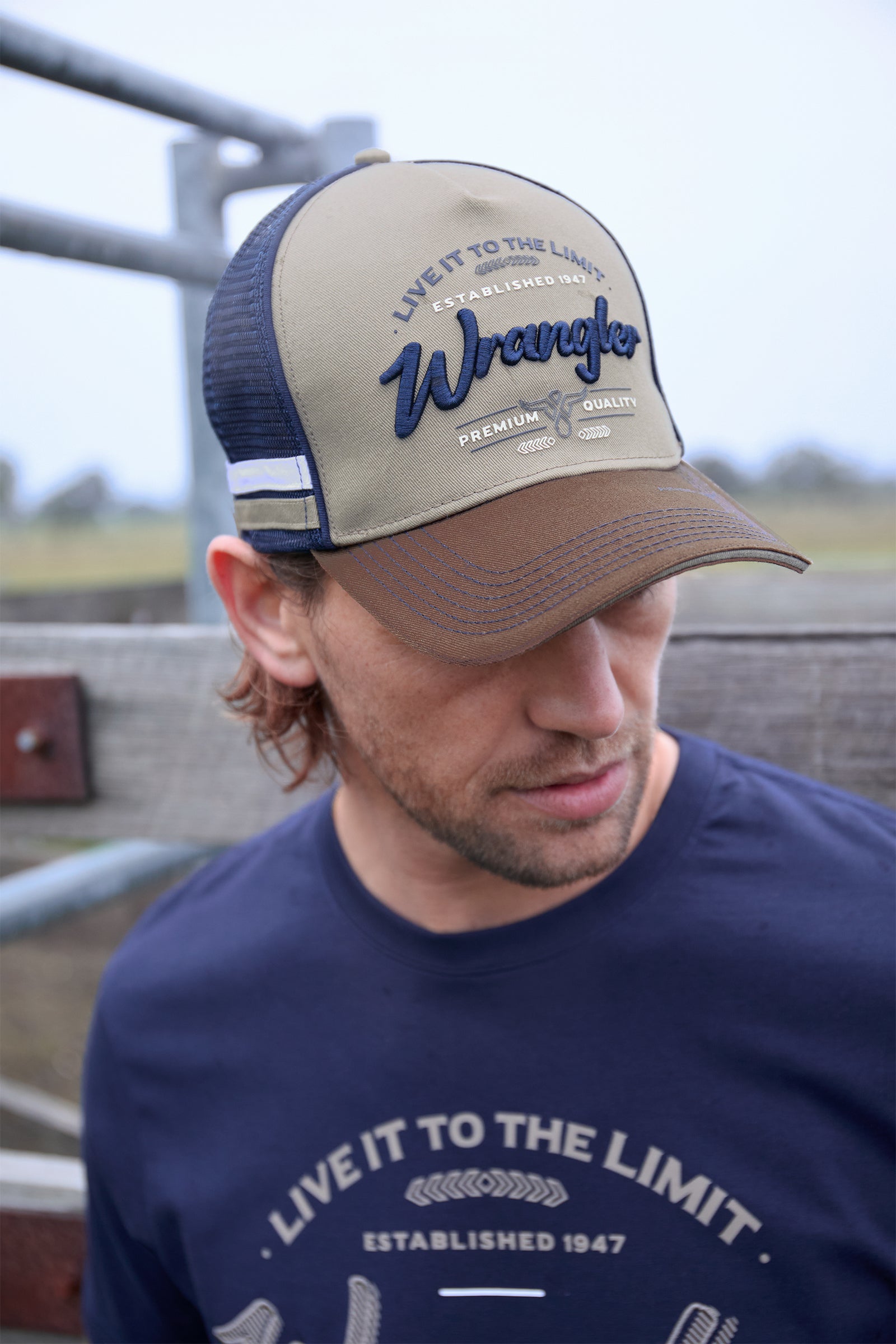 Man wearing a beige and brown cap with Wrangler branding, standing in an outdoor setting.