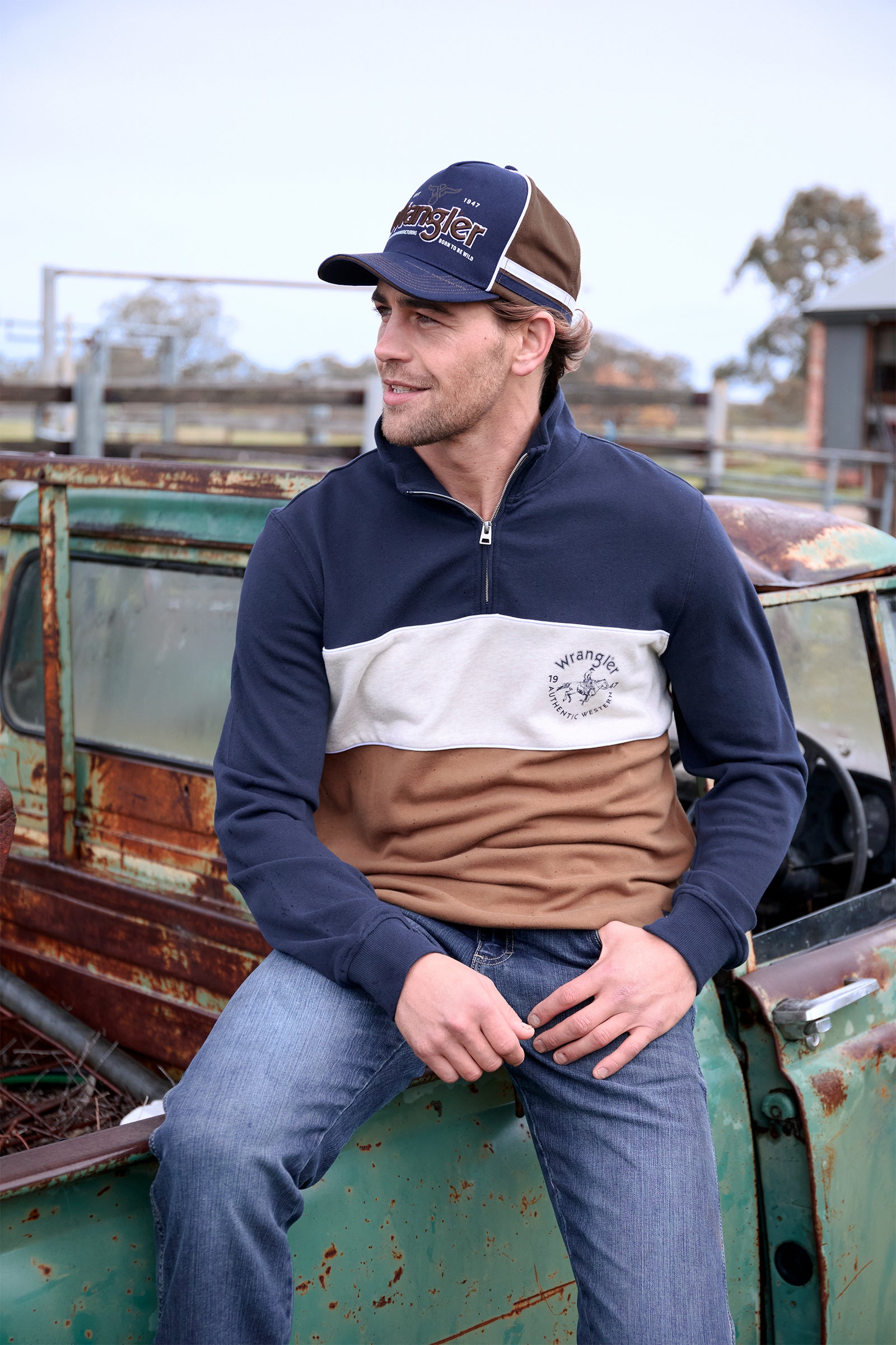 Man wearing a navy and brown pullover with a cap, sitting on an old truck.