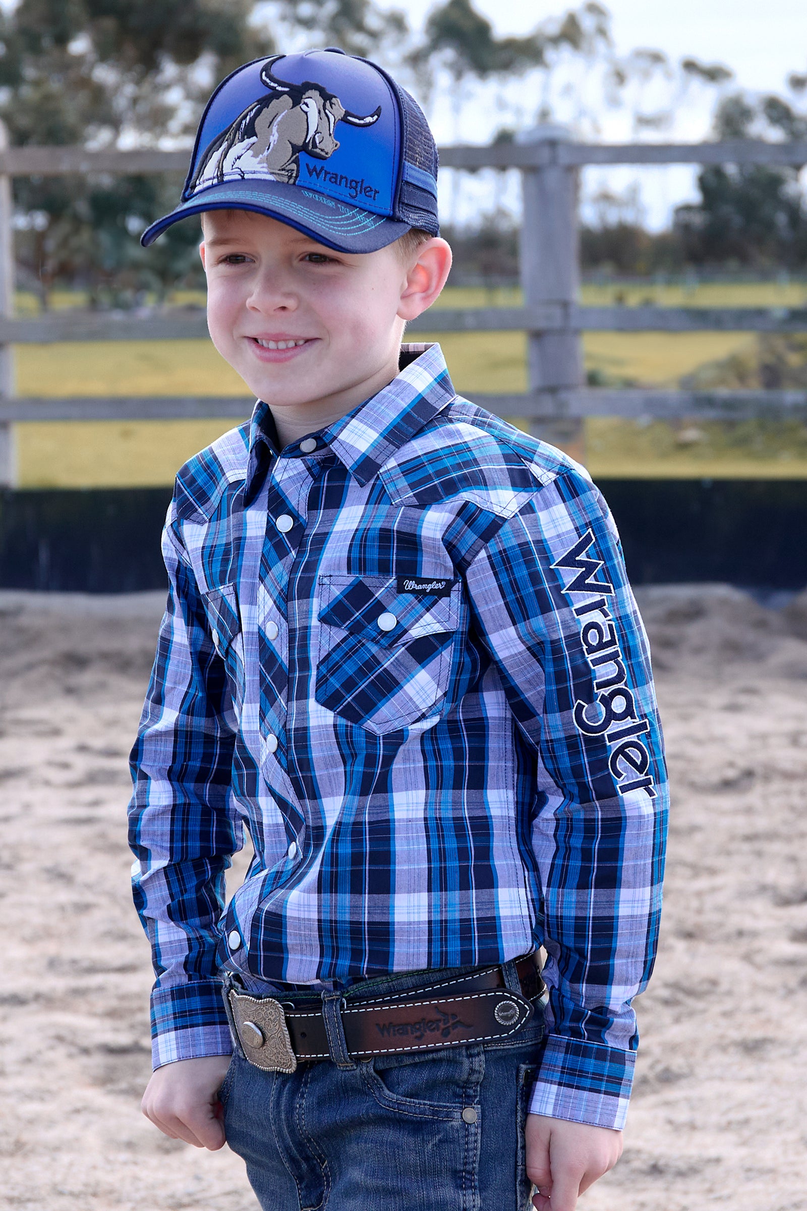 Young boy wearing a blue plaid shirt with a visible brand logo, standing in an outdoor setting.