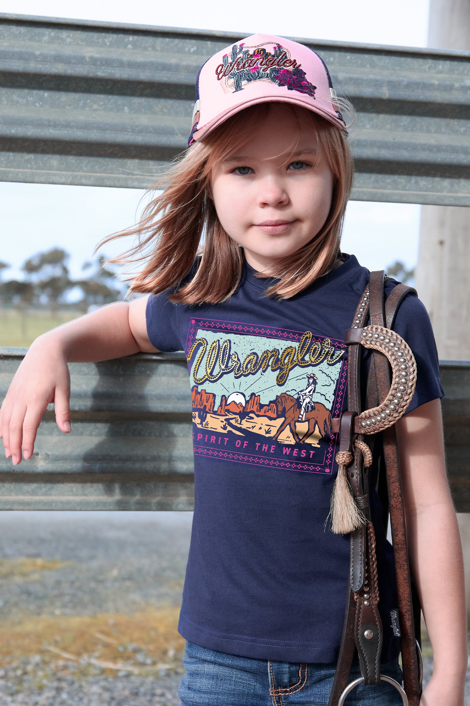Child wearing a Wrangler t-shirt and pink cap, standing outdoors.