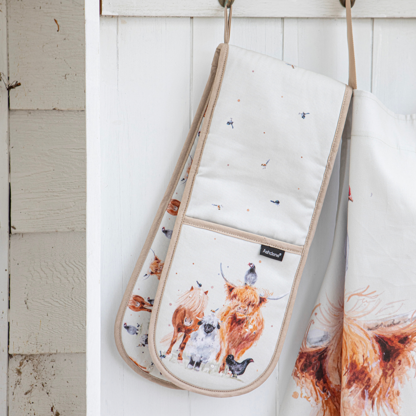 A cream-colored quilted kitchen towel or apron with a watercolor print of Highland cows, sheep, and birds, hanging on a white wooden peg against a rustic beige wall.