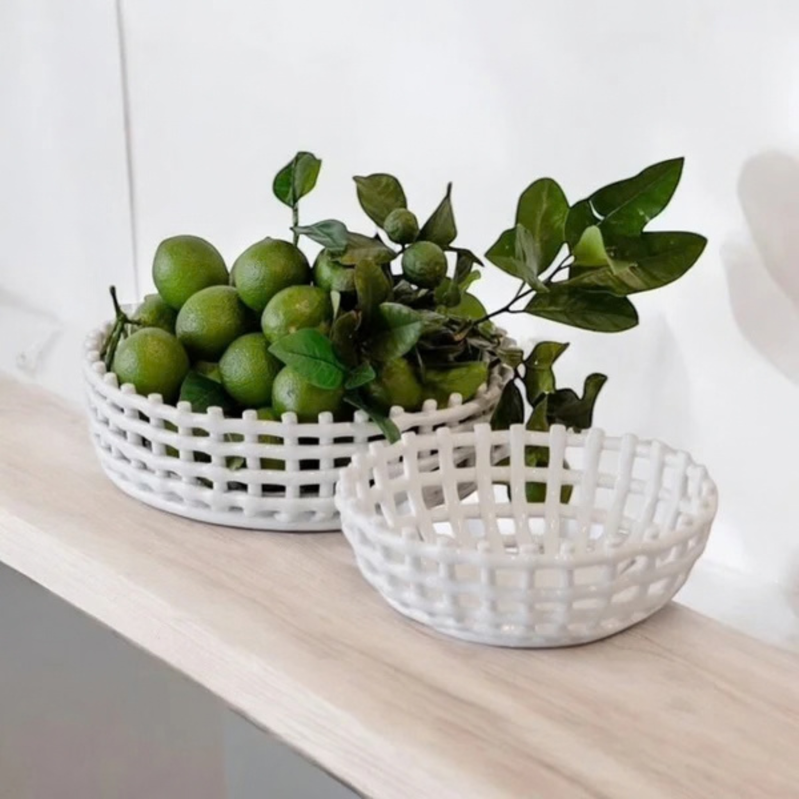 Two white plastic woven basket trays, one filled with green citrus fruits and a leafy branch and the other empty, displayed on a light wood shelf against a white wall.