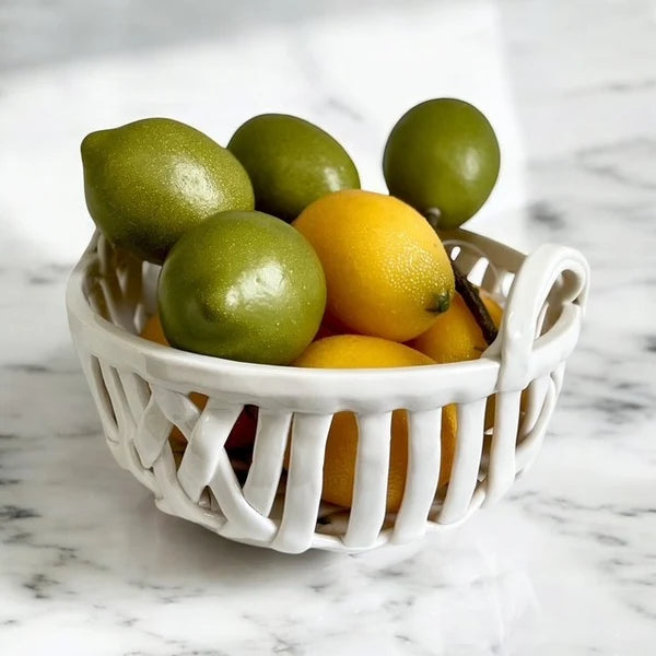 A white ceramic fruit basket filled with green and yellow lemons, set on a white marble surface.