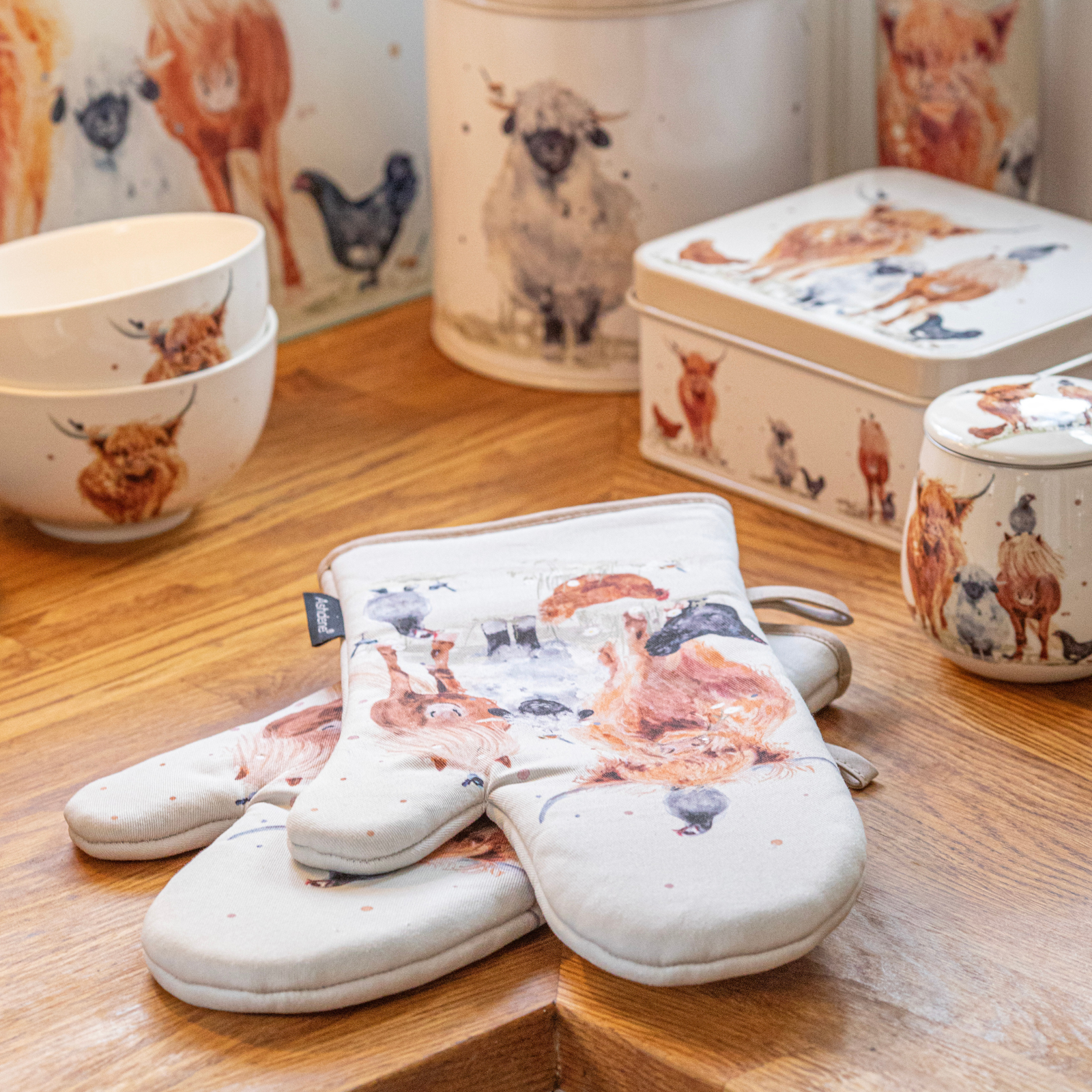 A pair of cream oven mitts with a rustic watercolor animal print, displayed on a wooden tabletop alongside matching ceramic canisters and bowls.