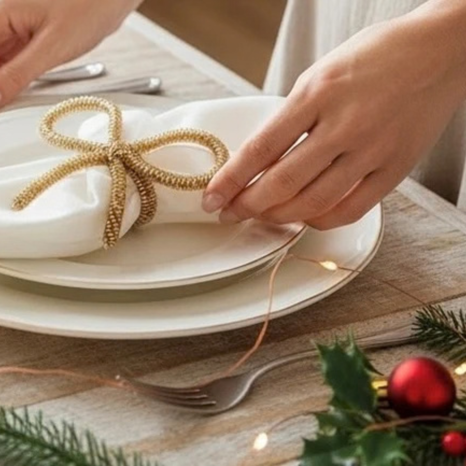 A close-up of hands placing a textured gold bow-shaped napkin ring on a white folded napkin atop stacked white plates, set on a wooden table with holiday greenery, a fork, and warm string lights.