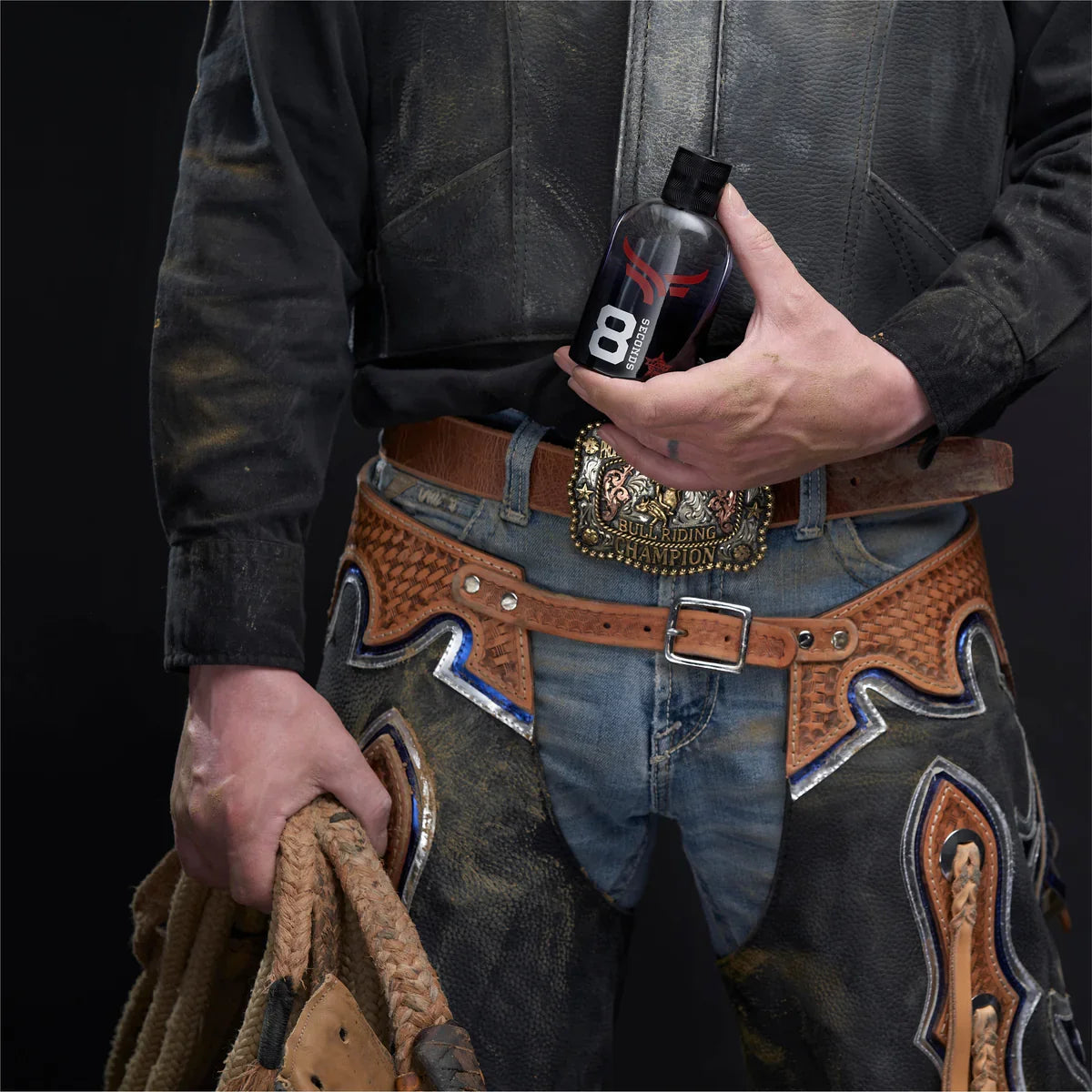 A close-up of a person wearing western-style leather chaps and a decorative belt, holding a black bottle labeled 8B in one hand and a rope in the other against a dark studio background.