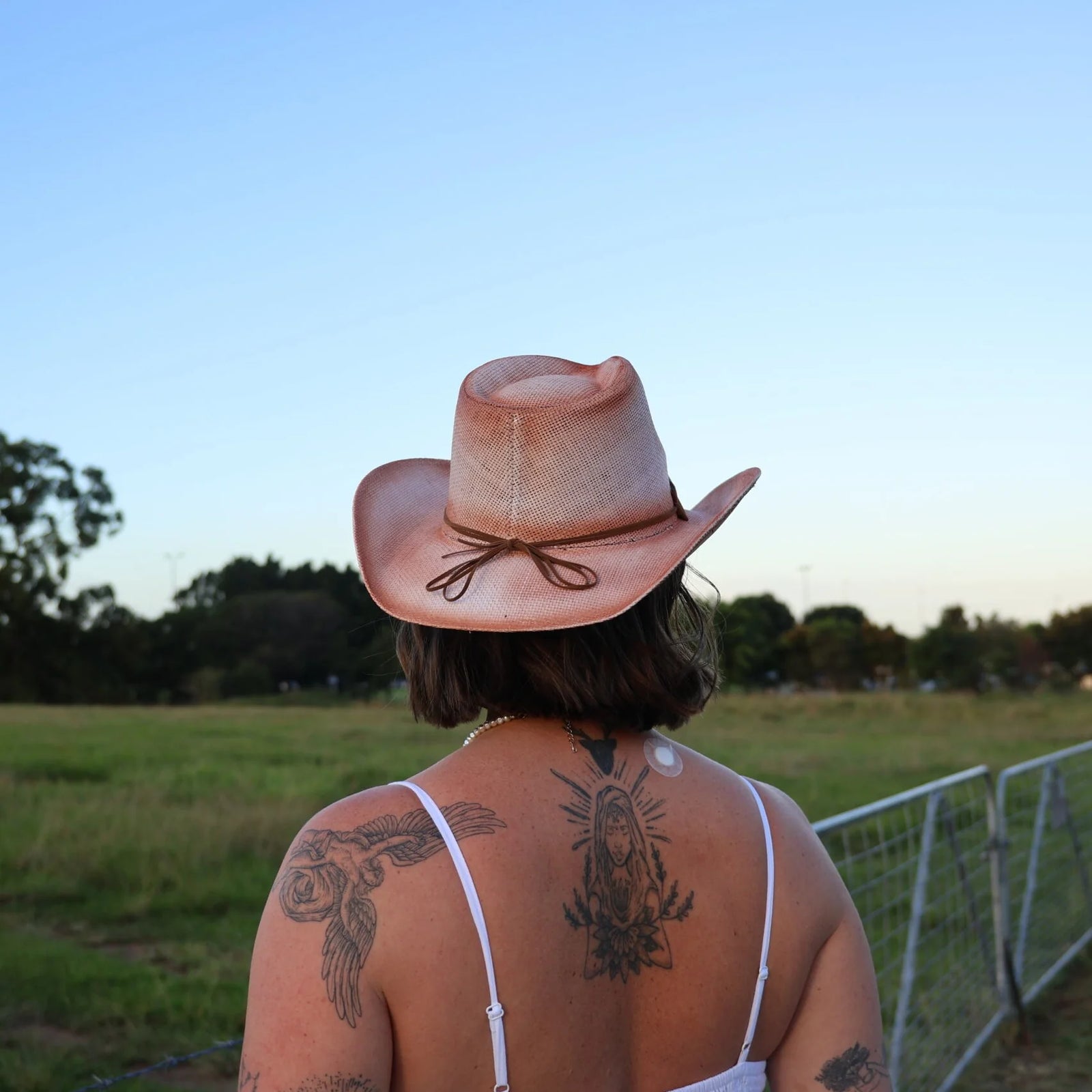 Person wearing a cowboy hat with tattoos on their back, standing in an open field.
