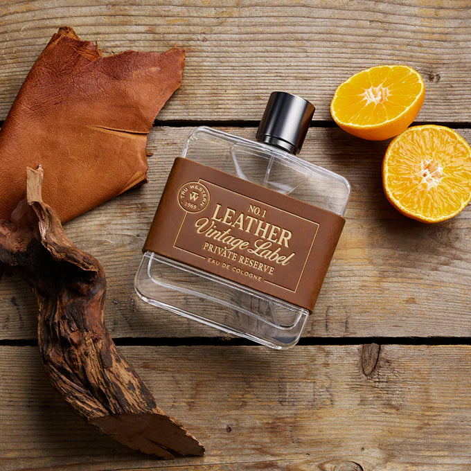 Tru Western Leather Vintage Private Reserve Label Eau De Cologne 100ml. Perfume bottle labeled 'Leather Vintage Label' on a wooden surface with orange halves and leather pieces.