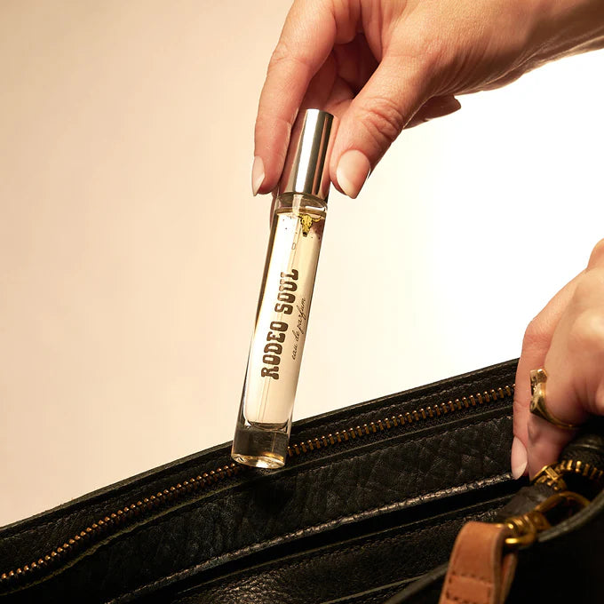 Hand holding a small perfume bottle labeled 'Rodeo Suds' over an open black handbag.