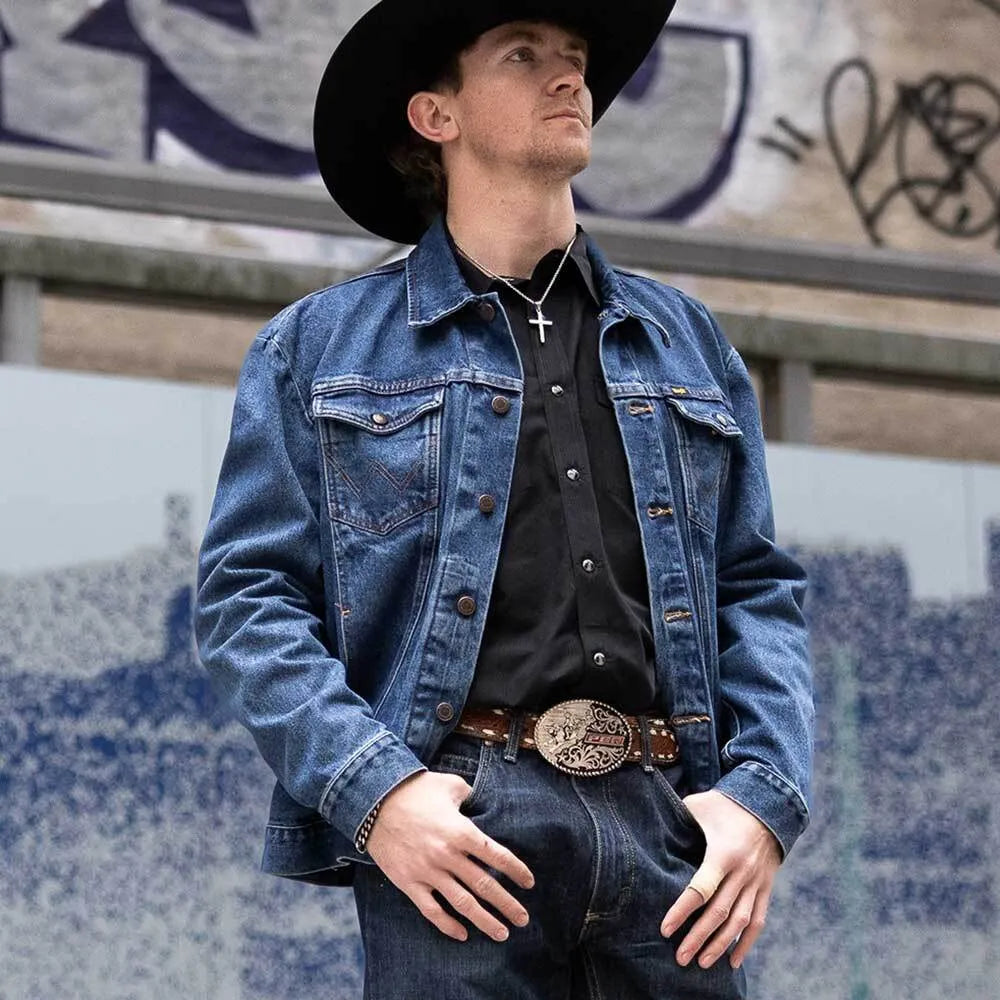 Man wearing a denim jacket and black hat in front of a graffiti-covered wall