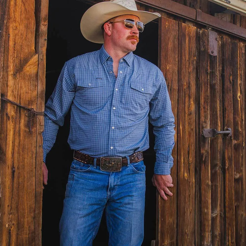 A man wearing a blue and white checkered long-sleeve western shirt, blue jeans, a brown leather belt with a large silver buckle, and a beige cowboy hat, standing in a rustic wooden doorway.