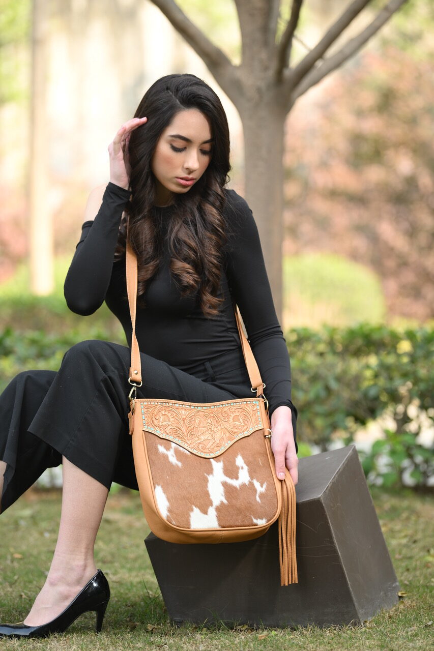 The Design Edge Women's Santiago Tooled & Hide Handbag. A woman sitting outdoors wearing a black outfit, holding a brown handbag with hand-carved 'lady on the horse' details and a leather tassel.