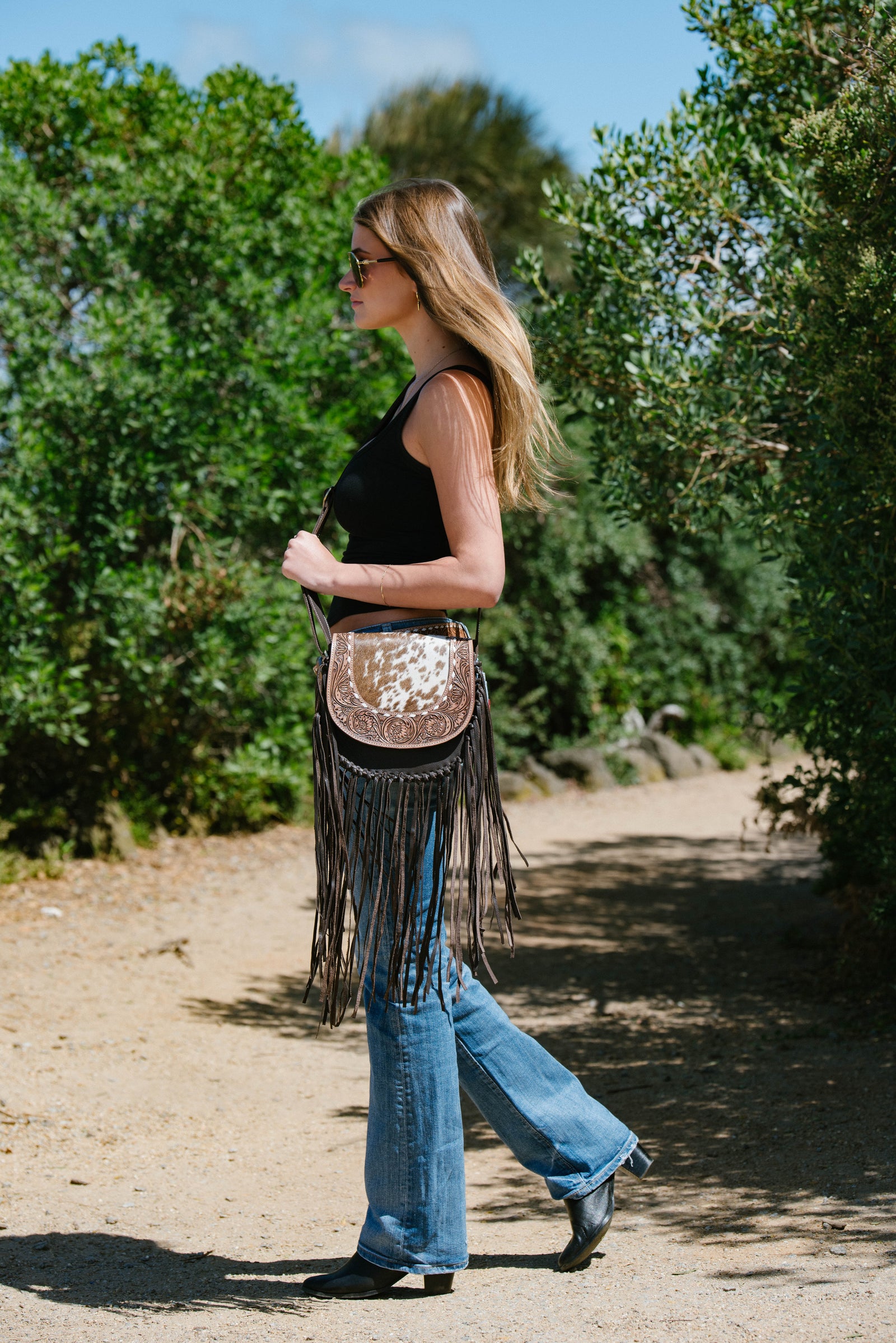 Woman walking outdoors wearing a black tank top, blue jeans, and a fringed handbag.
