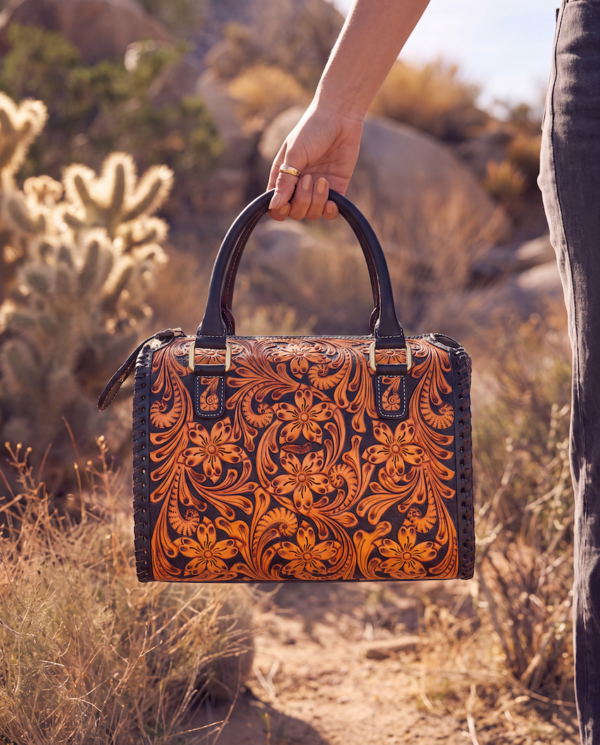 A hand holds a tooled leather satchel bag with a floral western pattern in orange and brown on a black background, set against a desert landscape background.