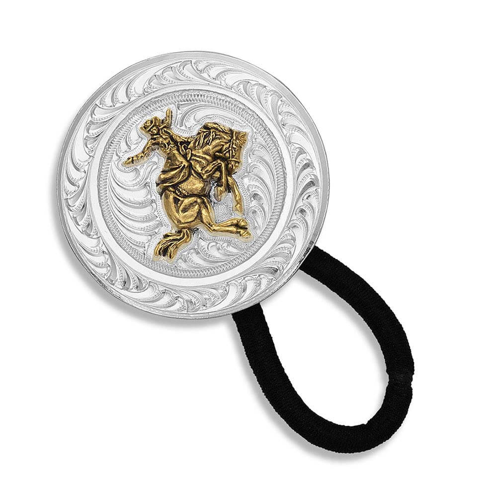 Montana Silversmiths Ponytail Holder. A round silver concho with an engraved design and an antiqued gold figure of a cowgirl on a bucking horse in the center, attached to a black ponytail holder with silver-toned tips.