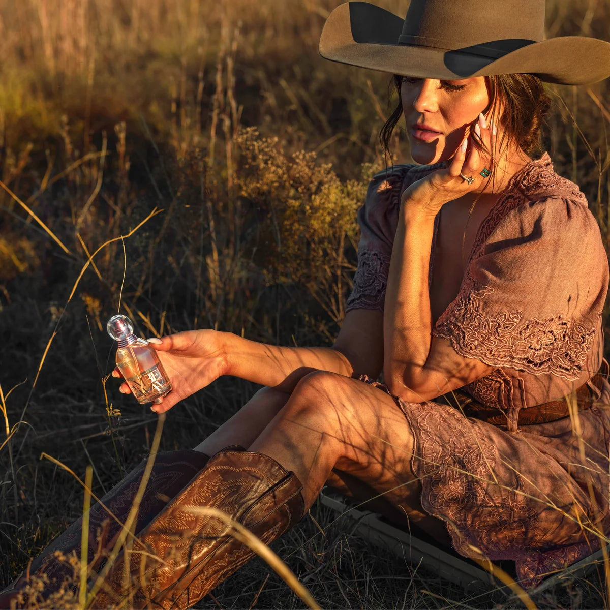 A woman in a cowboy hat and brown dress sits in a grassy field at golden hour, holding a small clear bottle with amber liquid. The scene is warmly lit with long shadows, and the product bottle is the focal point.