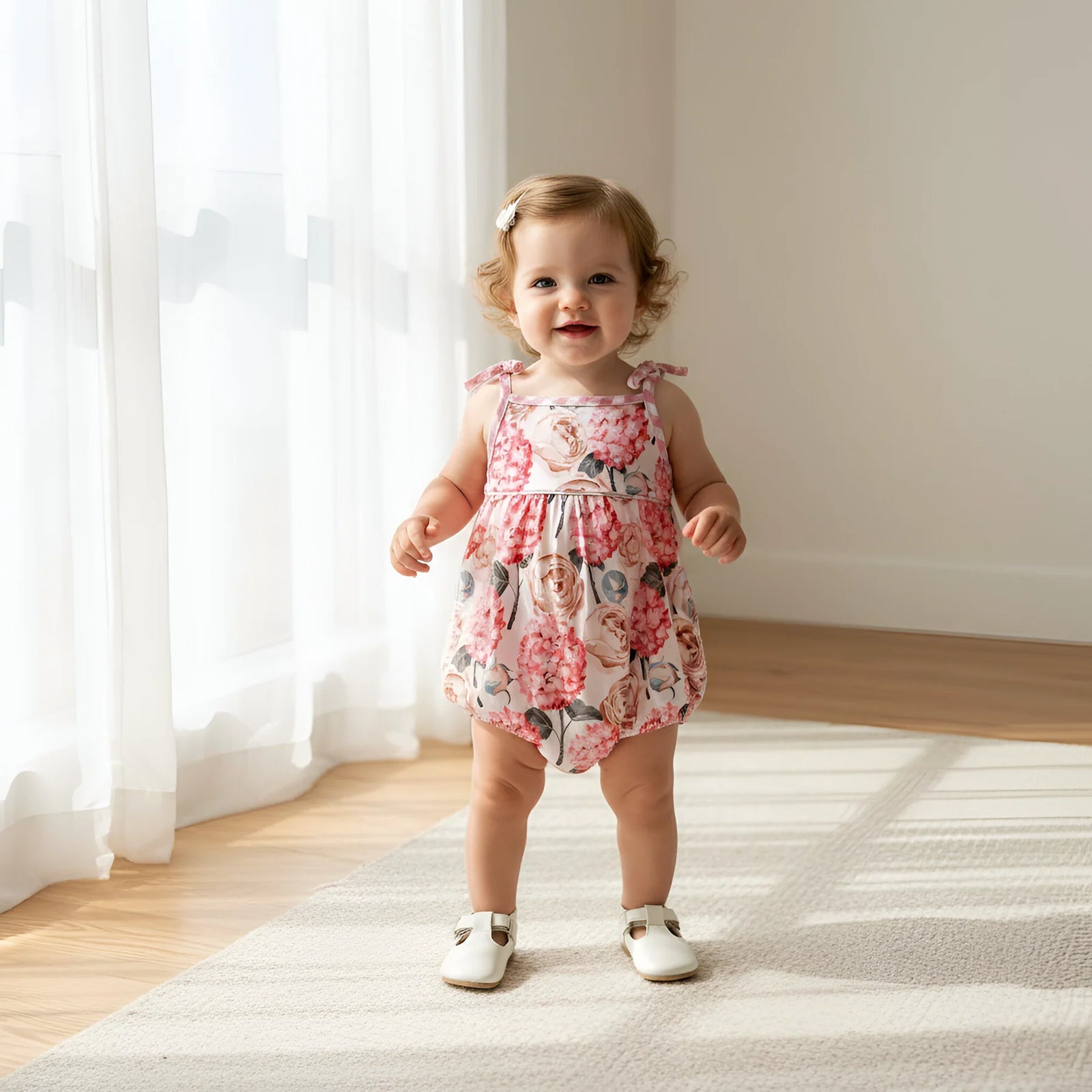 A toddler girl wearing a pink floral sleeveless romper and white shoes stands on a light rug in a bright room with sheer white curtains and wooden floors.